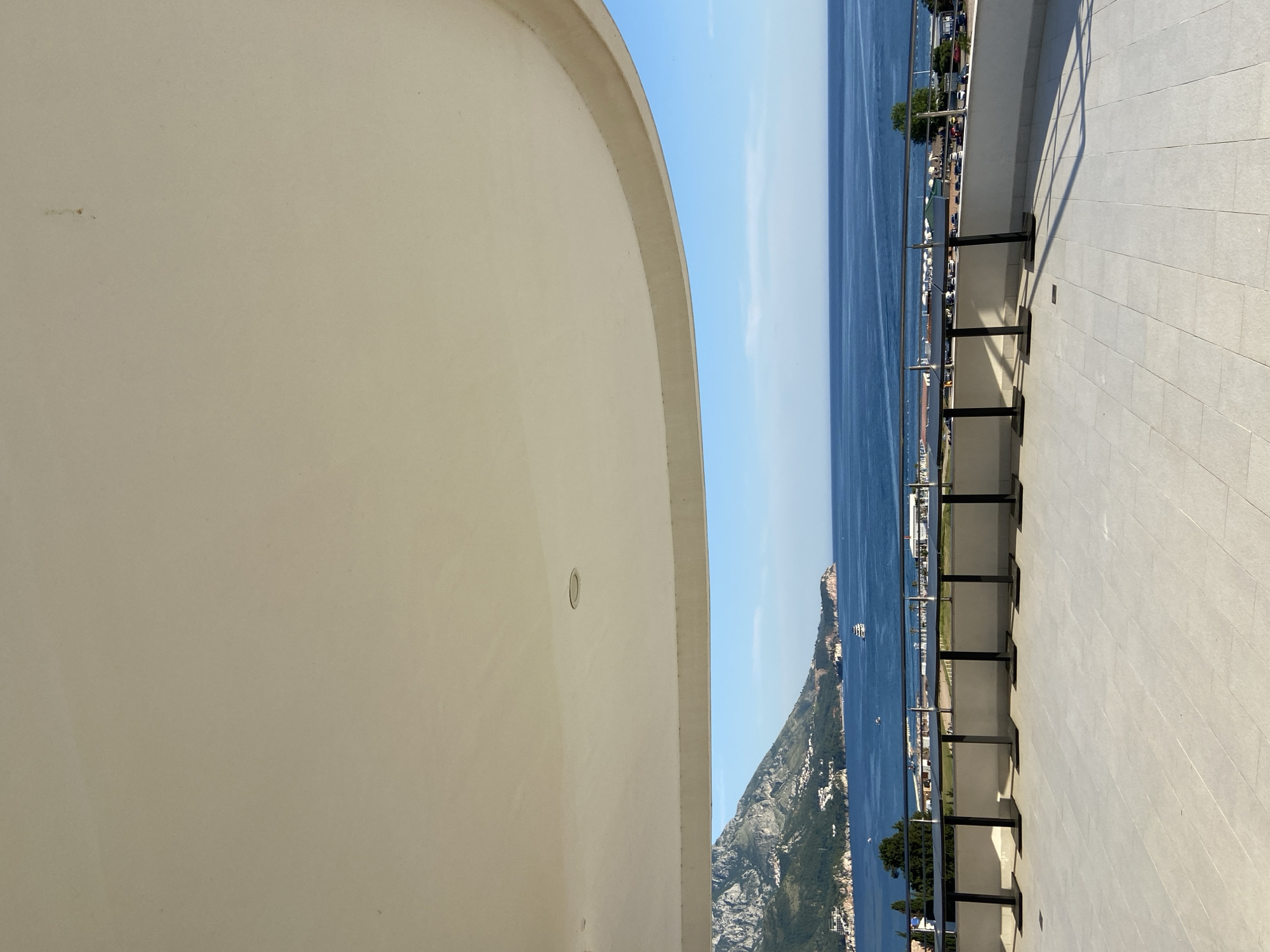 The image presents an exterior view from a balcony in Boreti, Montenegro. A large, rounded, white architectural element dominates the left side. To the right is a balcony with a modern railing consisting of glass panels and black supports. Beyond the railing is the Adriatic Sea, stretching towards the horizon. The coastline features a rugged mountain range covered with vegetation. The sea is dotted with small boats. Buildings and infrastructure are visible along the coastline. The sky is clear blue, with some faint clouds. The sun appears to be shining brightly, as indicated by the shadows on the balcony floor.