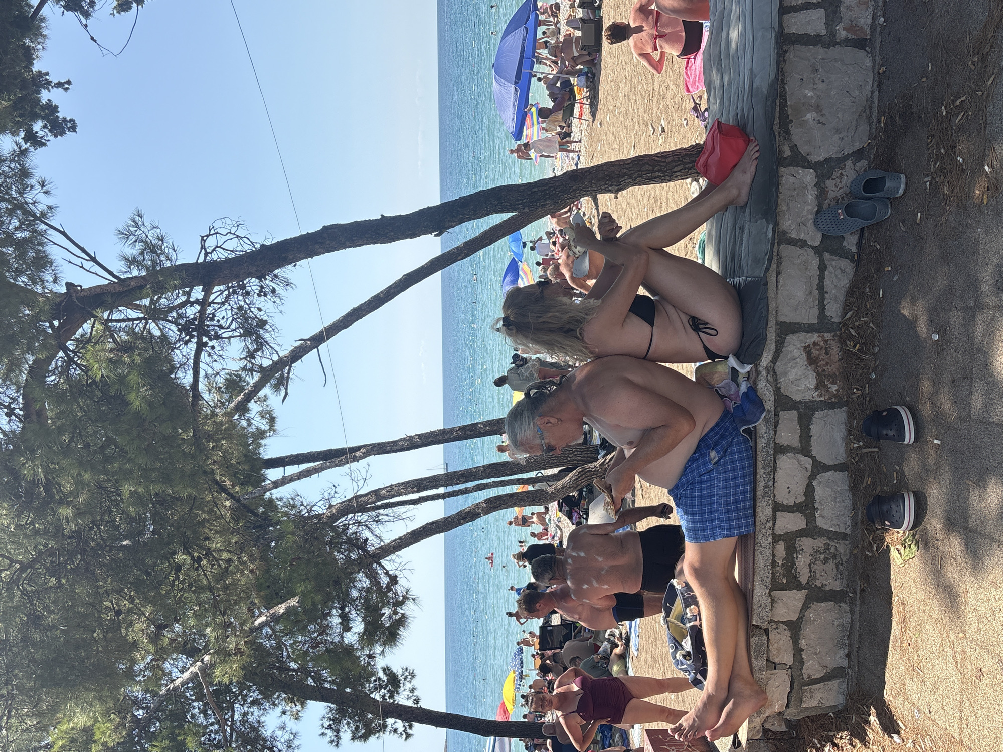 In Bar, Montenegro, on a sunny day, a beach scene unfolds under a canopy of pine trees. A stone wall, likely part of a seating area, runs horizontally across the foreground. Two individuals, a man and a woman, are seated on this wall, backs turned to the viewer. The man, wearing blue checkered shorts, is holding and looking at his phone. The woman, in a black bikini, is also engaged with her phone. Their feet are bare, resting on a dark mat. Beside them, on the sandy ground, are two pairs of casual shoes.
Beyond the wall, the beach is populated with numerous people enjoying the sun and sea. Beach umbrellas, in blue and yellow, dot the sand. Some individuals are standing, while others are seated or lying on towels. The turquoise water of the Adriatic Sea stretches to the horizon, where a sailboat and a few distant figures can be seen. The sky is clear and blue. The trees in the foreground cast shadows on the sand, indicating a bright, sunny atmosphere. Other beachgoers, partially obscured by the trees, are visible in the mid-ground and background, some swimming or wading in the water, others relaxing on the shore.
