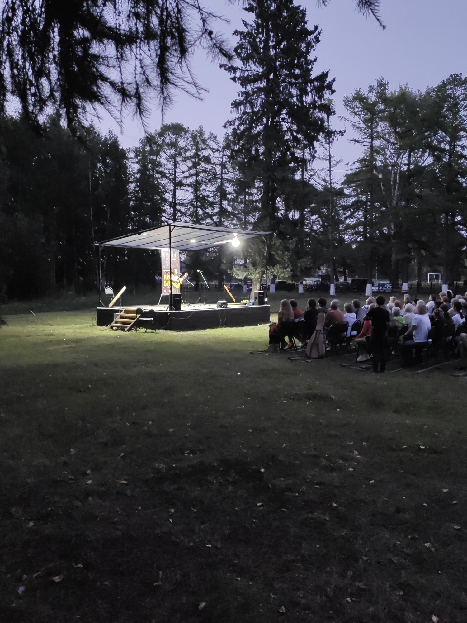 An outdoor evening concert is taking place on a grass field in Yurino, Russia. A stage, covered by a translucent roof and illuminated by spotlights, is set up in the background. A lone musician, wearing a hat and playing a guitar, is performing on stage. An audience of approximately 50 people is seated in chairs facing the stage. The surrounding environment includes trees and a dimly lit sky, suggesting twilight or early night.