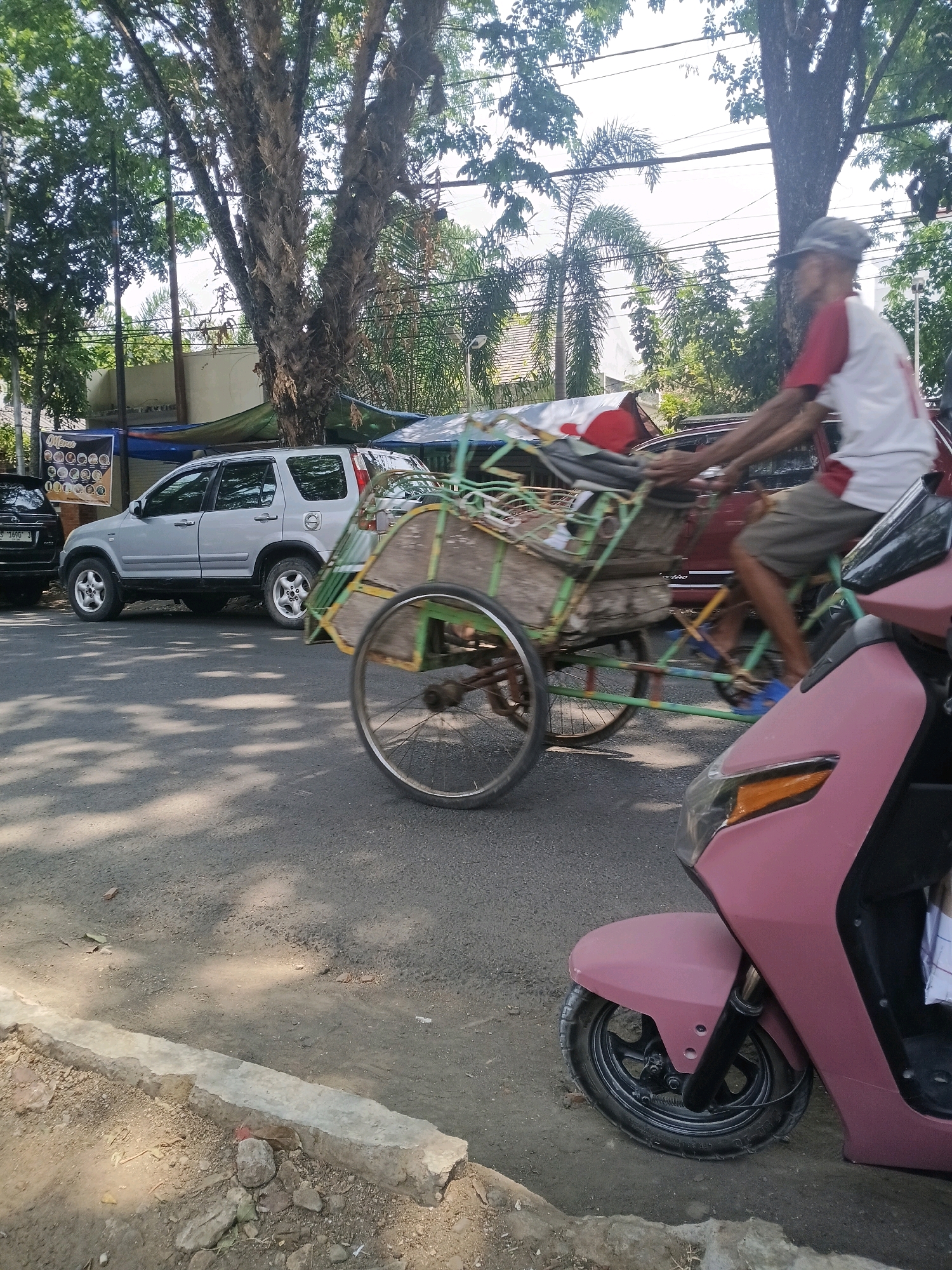 In Purwodadi, Indonesia, a man is pedaling a pedicab loaded with goods down a sun-dappled asphalt road. The pedicab, painted green and yellow, has a wooden cargo box. Behind the pedicab, a silver SUV is parked. In the foreground, the front wheel and fender of a pink scooter are visible. Large trees with sparse foliage line the street, casting shadows on the road. The sky is bright and clear.