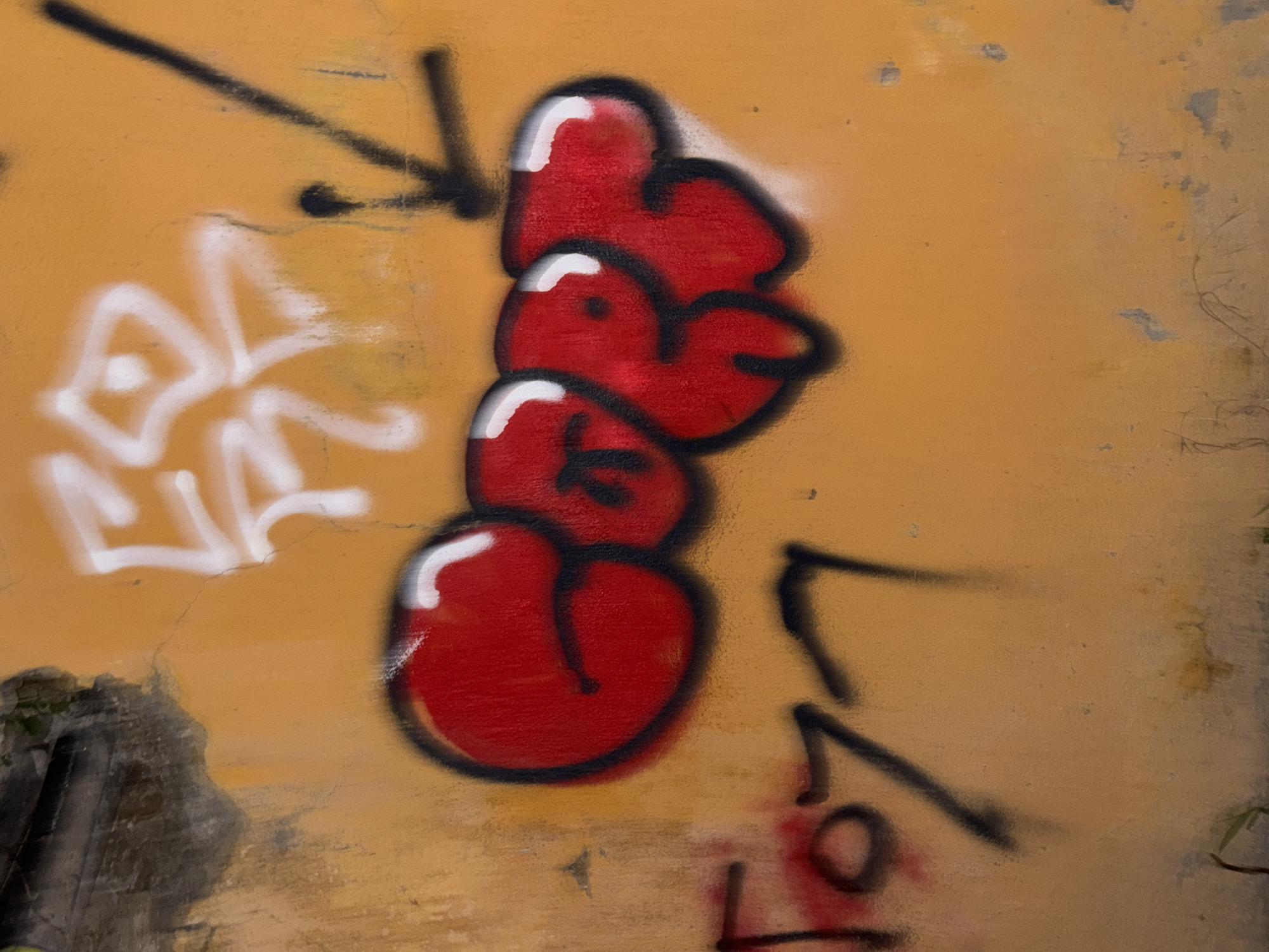 The image is a close-up, eye-level, outdoor shot of graffiti on an orange, textured wall in Unknown, Unknown. The wall shows signs of wear and tear, with cracks and some darker patches near the top left. The graffiti features several distinct elements. In the upper section, there are white spray-painted characters that are abstract and somewhat stylized, with a diagonal black line extending from the right of these characters. Below these, a prominent red graffiti tag with a black outline and white highlights fills the central part of the image. This tag appears to be in a bubble letter style and possibly spells out "EGRF" or a similar word. To the left and slightly below the red tag, there is another piece of graffiti in black and red, which appears to be numbers or a tag, possibly "1077". The overall lighting suggests it is daytime, with some shadows cast by the graffiti. The lower portion of the image shows the continuation of the orange wall and some faint, abstract lines or scratches.