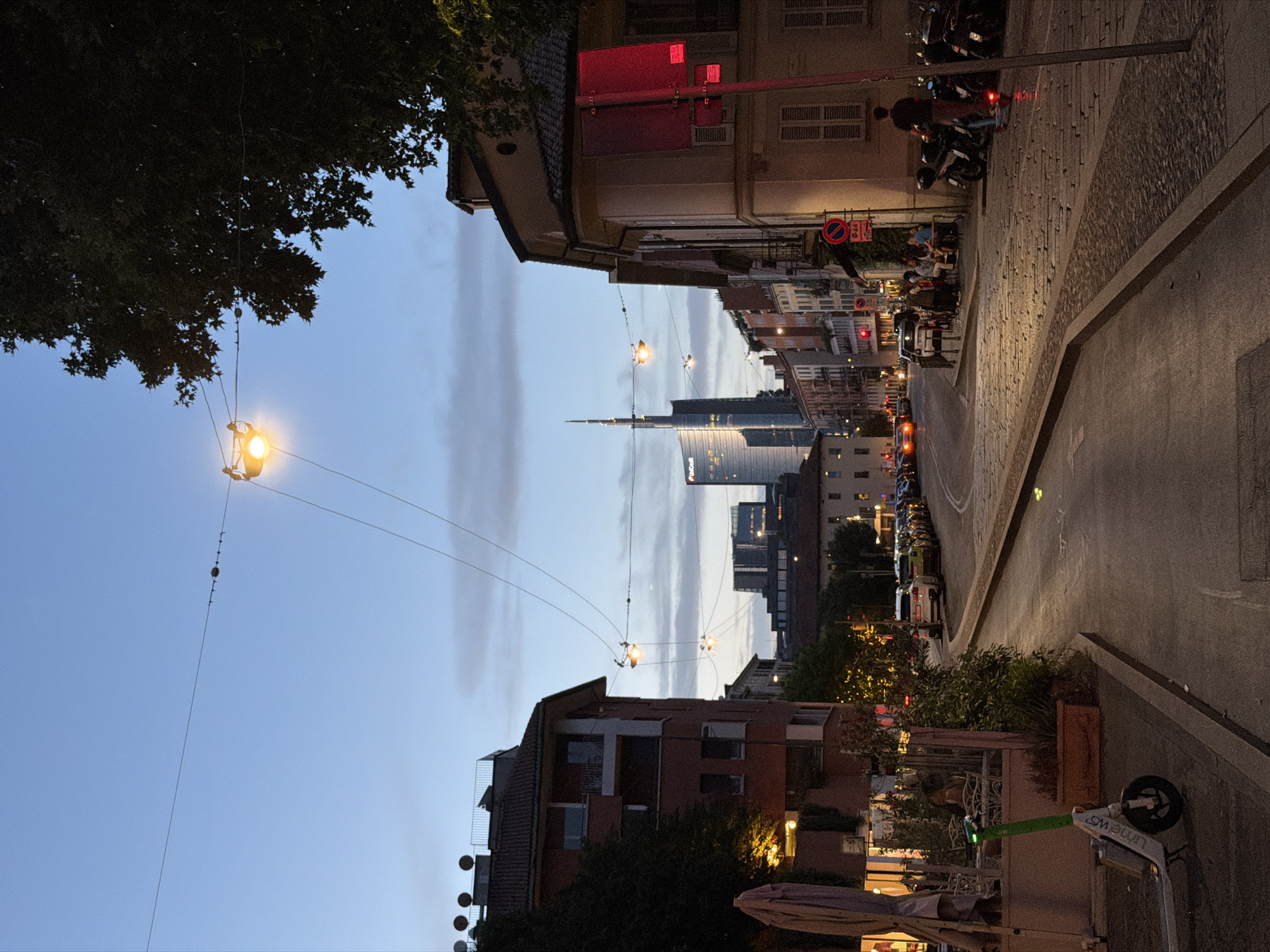 This image captures a twilight street scene in Milan, Italy, specifically in the Porta Nuova district, as evidenced by the prominent Unicredit Tower in the background. The perspective looks down a cobblestone street lined with buildings on both sides. Overhead, streetlights are strung across wires, casting a warm glow.
On the left side of the street, a red apartment building with balconies stands. In front of it, a white electric scooter with green accents is parked on the sidewalk. Further down, outdoor seating with tables and chairs for a cafe or restaurant is visible.
On the right side of the street, a cream-colored building with shuttered windows is featured. A bright red sign, likely a traffic or business sign, is attached to a pole. A person is standing next to a row of parked motorcycles, some of which are visible.
The street itself is paved with cobblestones and shows a gentle curve. Cars are parked along the right side of the street and are driving on the road. The sky is a dusky blue with scattered clouds, indicating the transition from day to night. In the distance, the modern architecture of the Unicredit Tower, a skyscraper, stands out against the softer hues of the older buildings. The overall impression is a lively urban evening.