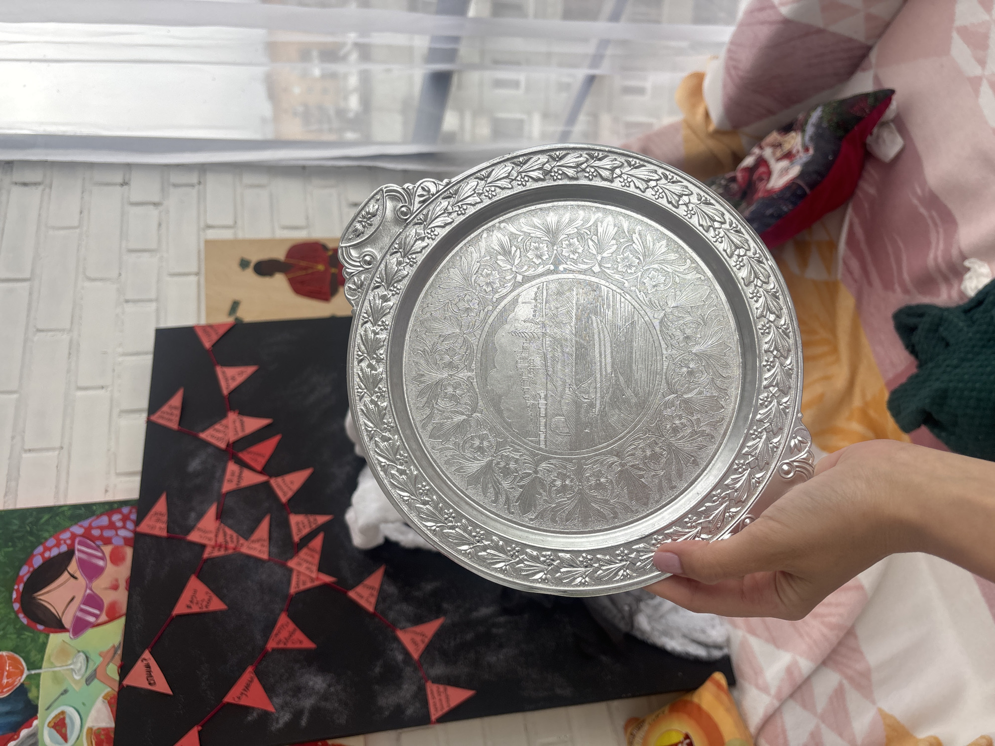A hand holds a silver-colored ornate serving tray with a detailed floral and architectural design. The tray is positioned above a scene including an illustration of a woman in sunglasses, a black surface adorned with red triangle flags strung together, and a pillow. The background features a white brick-like wall with a window partially visible. The scene is indoors. There is no information about the city of Krasnoyarsk, Russia in the image.