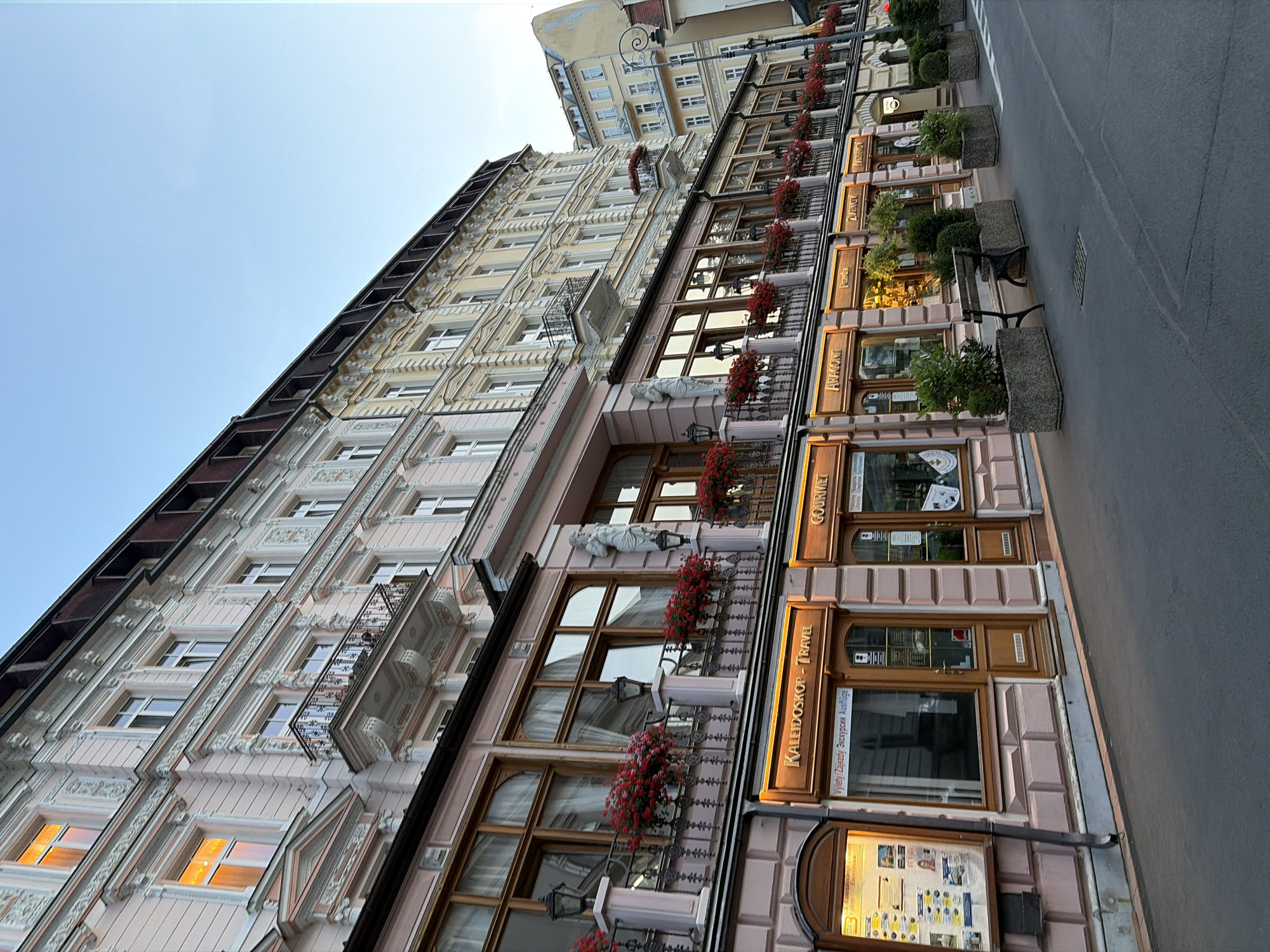The image depicts a street scene in Karlovy Vary, Czechia, focusing on a row of ornate, multi-story buildings lining a paved street. The architecture is classic European, with light pink and pale yellow facades, detailed stucco work, and ornate balconies adorned with red flowers. Several businesses with wooden-framed windows and signage are visible at street level, including "KALEIDOSKOP - TRAVEL" and "GOURMET." The street is empty of people, with the exception of a single bench situated next to planter boxes containing greenery. The sky above is a clear, pale blue, suggesting it is either early morning or late afternoon. The overall impression is one of a well-maintained, historic urban environment.