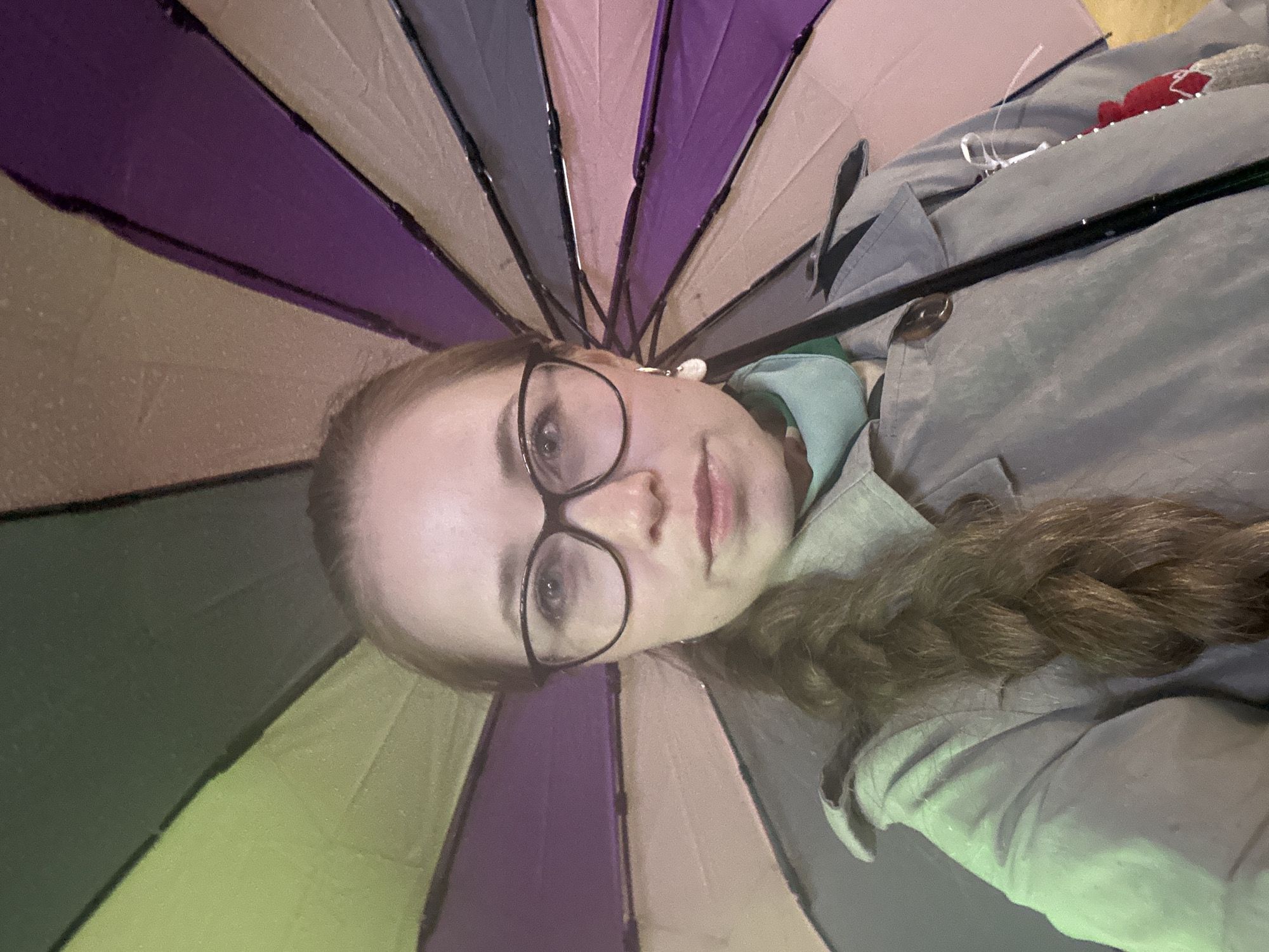 A woman with light skin, wearing glasses and a grey coat, is centered in the image, holding a colorful umbrella above her head. The umbrella has segments of purple, tan, grey, and pink. The background is dark, suggesting it is either night or a very overcast day. There are no clear indications of the location being Saint Petersburg, Russia, so it cannot be confirmed from the image alone.