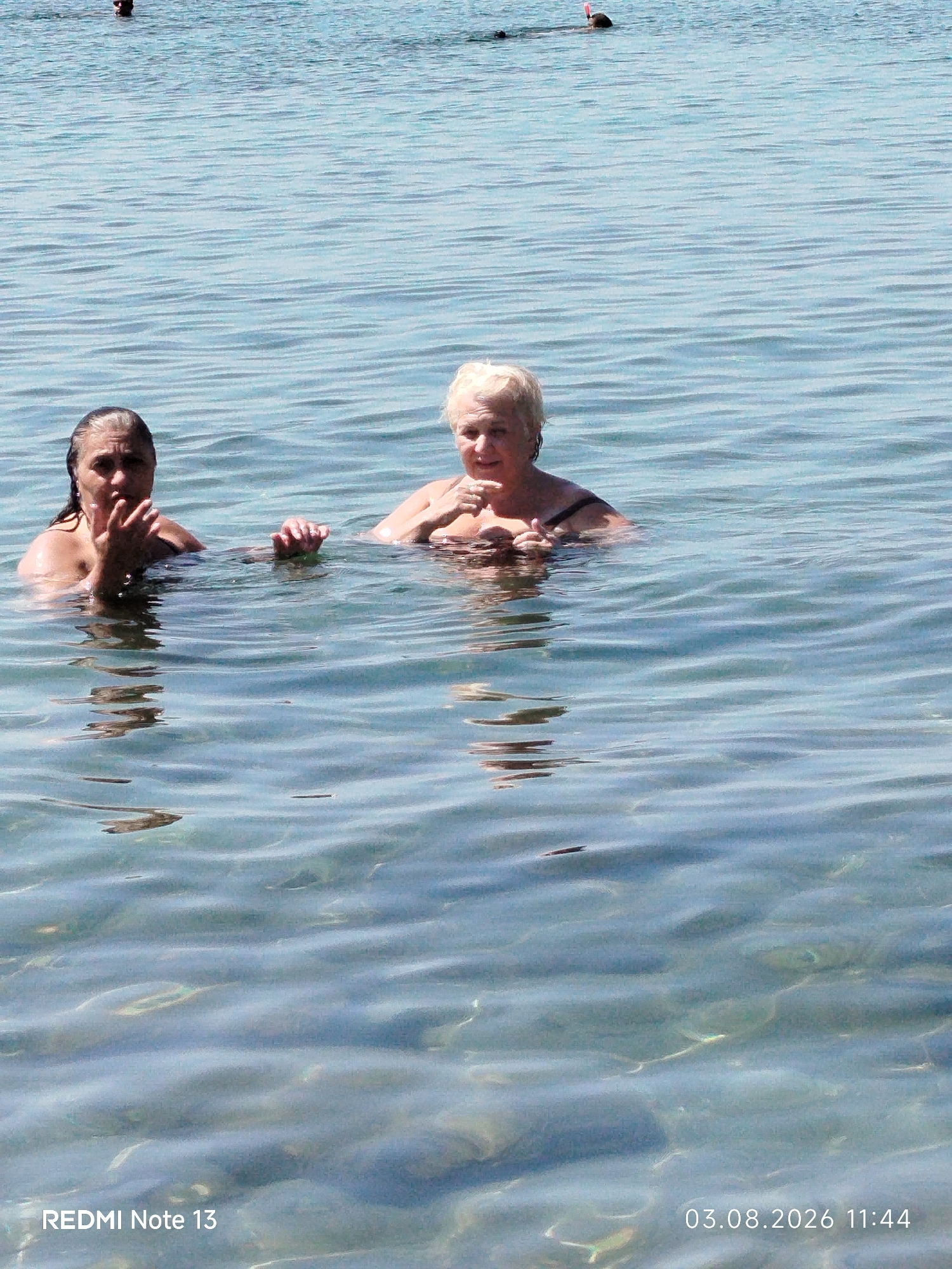 The image shows two women swimming in the clear blue waters of the sea. The woman on the left has dark, wet hair and is looking towards the camera with her hand raised. The woman on the right has blonde hair and is also looking towards the camera. In the background, two other individuals are swimming or snorkeling. The water is calm with visible ripples and sunlight reflecting off the surface. The image appears to have been taken from a low angle, looking out over the water. The timestamp "03.08.2026 11:44" is visible in the bottom right corner, and "REDMI Note 13" in the bottom left. The general setting is a coastal area, consistent with the beaches found in Sithonia, Greece, a region known for its clear waters and scenic coastline.