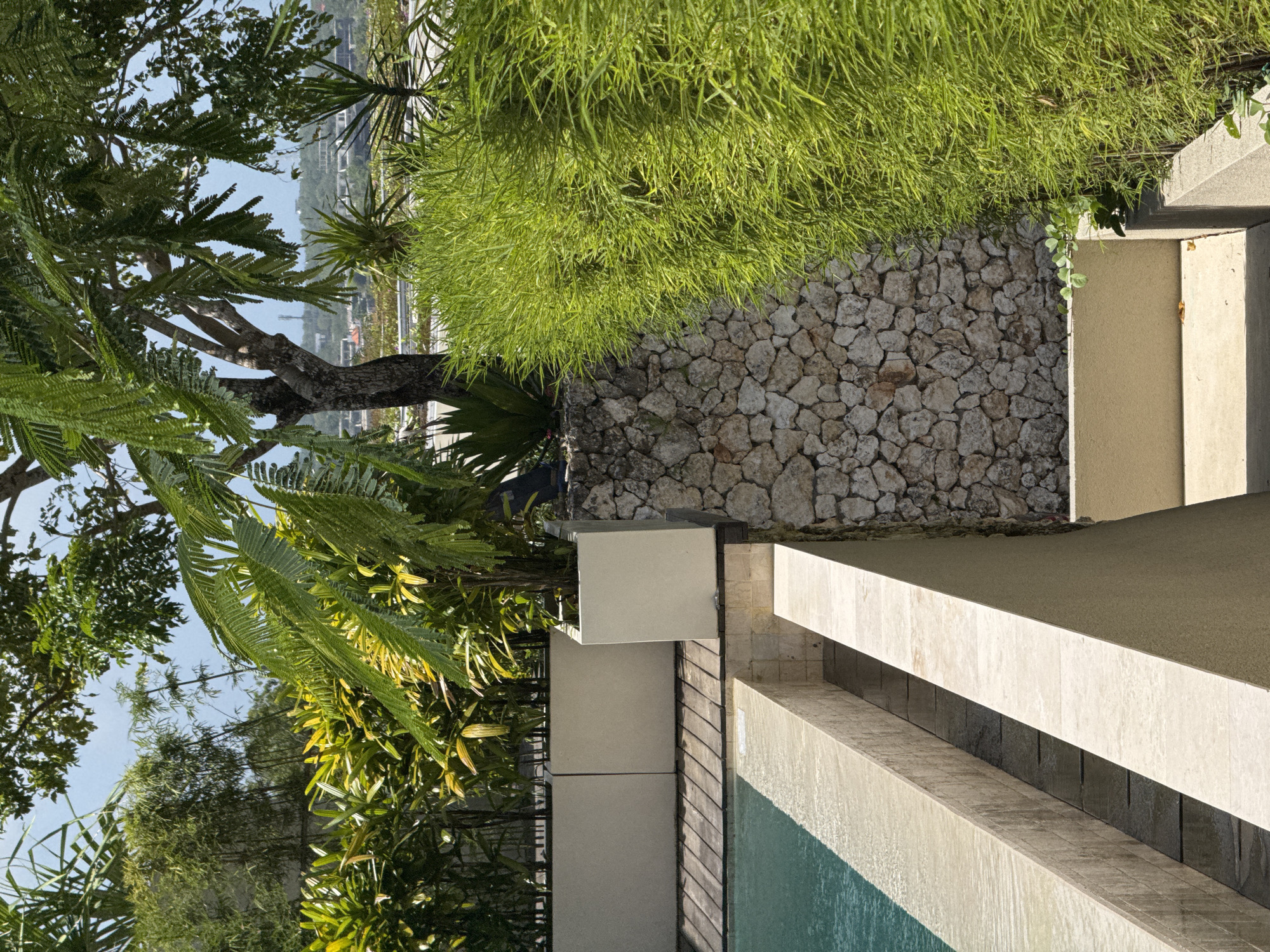 The image depicts a section of a property in Kuta Selatan, Indonesia, focusing on the landscaping and architectural elements near what appears to be a swimming pool. In the foreground, there's a partially visible pool with a light blue water surface, framed by light-colored stone or concrete structures and dark wood panels. Adjacent to the pool, a low wall or border extends. Behind this feature, a dry-stacked stone wall separates the pool area from an elevated patch of dense, green grass. The foliage includes diverse trees and plants, some native to tropical climates, partially obscuring the background. In the distance, some residential or commercial buildings are visible against a light blue sky.