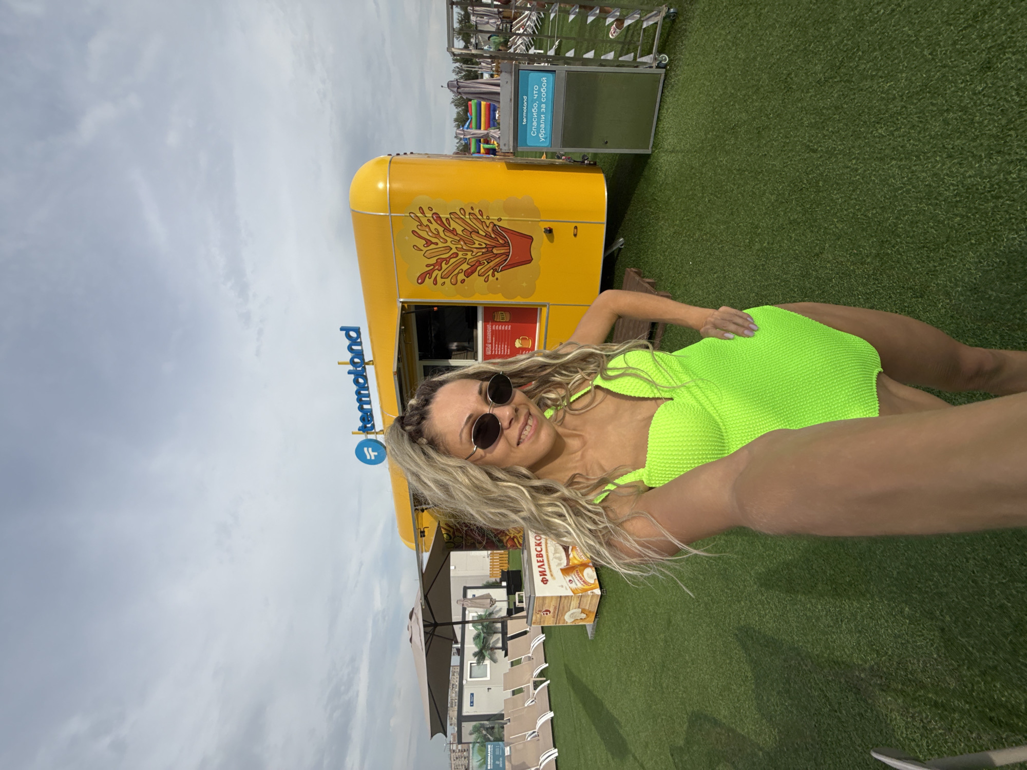 A woman with blonde, wavy hair and sunglasses takes a selfie outdoors. She is wearing a bright neon green one-piece swimsuit. Behind her, a yellow food truck with a logo that reads "termoland" is visible. The food truck has an illustration of french fries on its side. To the left of the food truck, there are lounge chairs and an umbrella. To the right, there is a trash receptacle with text in Russian. The scene appears to be a recreational area with artificial turf covering the ground. The sky is overcast with clouds. This location is identified as Rusanovo, Russia.