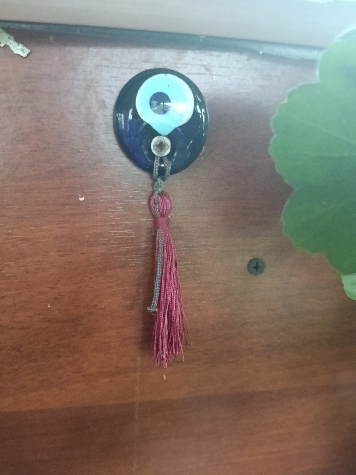 A close-up shot reveals a dark blue glass evil eye amulet with a light blue center. A maroon tassel with gray accents hangs from the amulet. The amulet is mounted on a brown wooden surface. A green leaf of a plant is visible to the right. A screw is also visible on the wood.
The location is not discernible from the image.