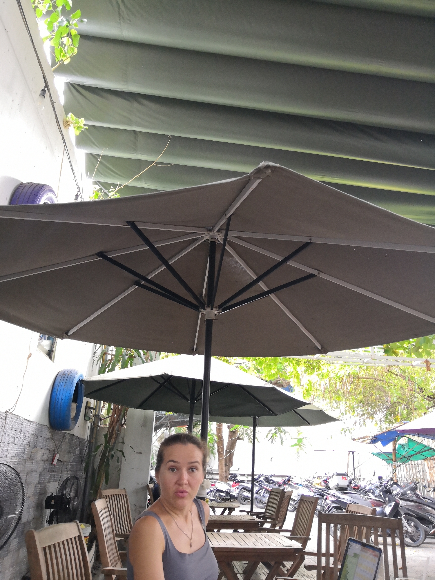 A woman in a gray tank top is sitting at a table outdoors under a large umbrella. She is looking at the camera with a surprised expression. In the background, there are several motorcycles parked, as well as other tables and chairs. The location is likely a cafe or restaurant in Nha Trang Ward, Vietnam, given the presence of motorcycles, outdoor seating, and tropical foliage.