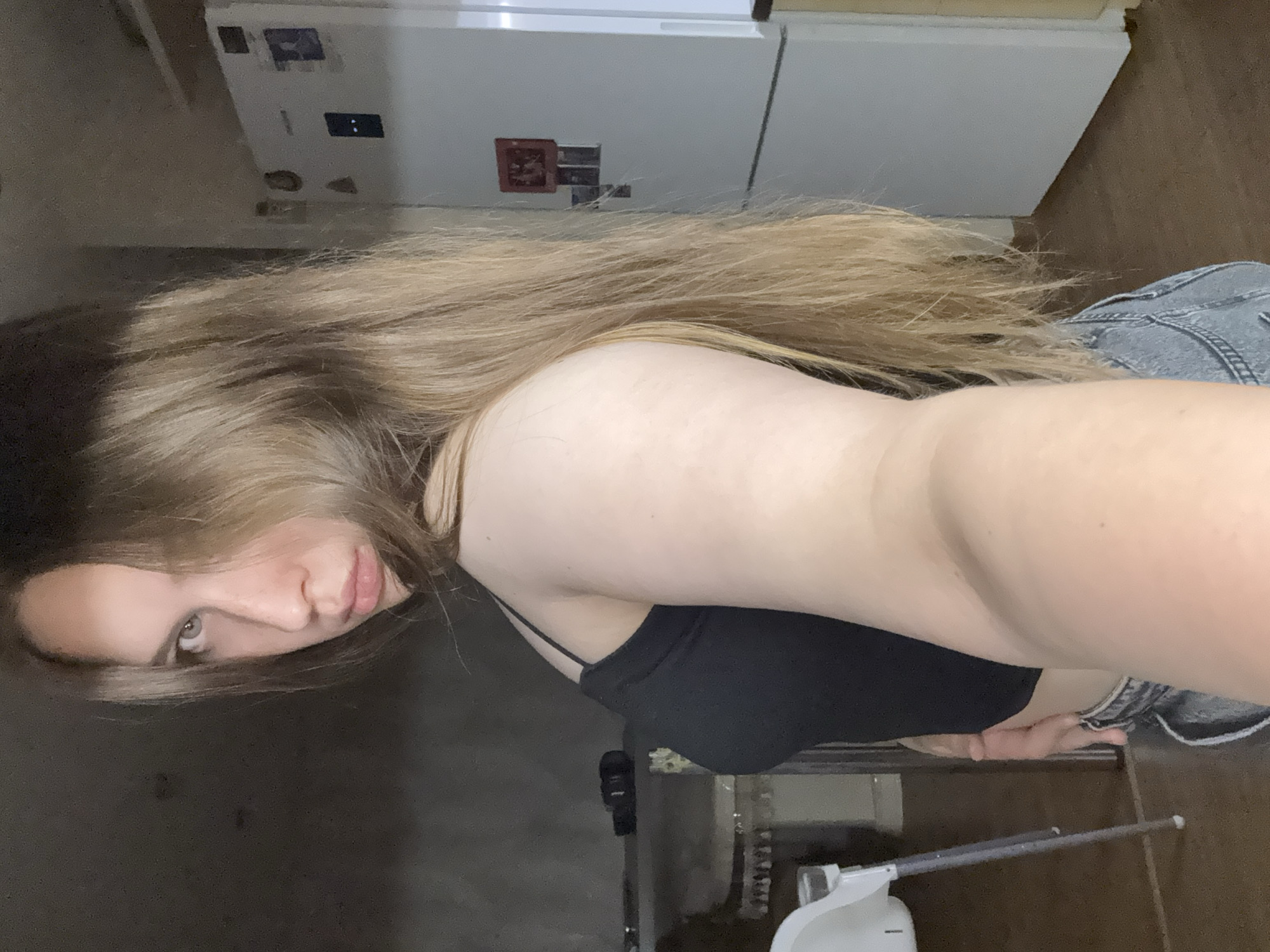 The image captures a selfie taken indoors in what appears to be a kitchen setting in Chaikovsky, Russia. The main subject is a person, likely female, with long, flowing light brown hair that cascades down her back and across her shoulder. She is wearing a black spaghetti-strap top, possibly a crop top or sports bra, and light-wash denim jeans or shorts. Her head is tilted, with her face partially visible, showing one eye, part of her nose, and slightly parted lips, conveying a neutral or subtly contemplative expression.
In the background, a large white double-door refrigerator-freezer unit is prominent. Several small magnets are attached to its doors; one on the left door appears to be a small picture frame magnet with a reddish image, and faint dark symbols or lights are visible on its control panel. The floor is a warm brown, resembling wood or laminate. To the bottom right, a white steam cleaner or similar household appliance with an extending handle is visible on the floor. The lighting in the room is somewhat dim, suggesting an indoor shot not taken in bright daylight. The image is oriented sideways, characteristic of a casual selfie.