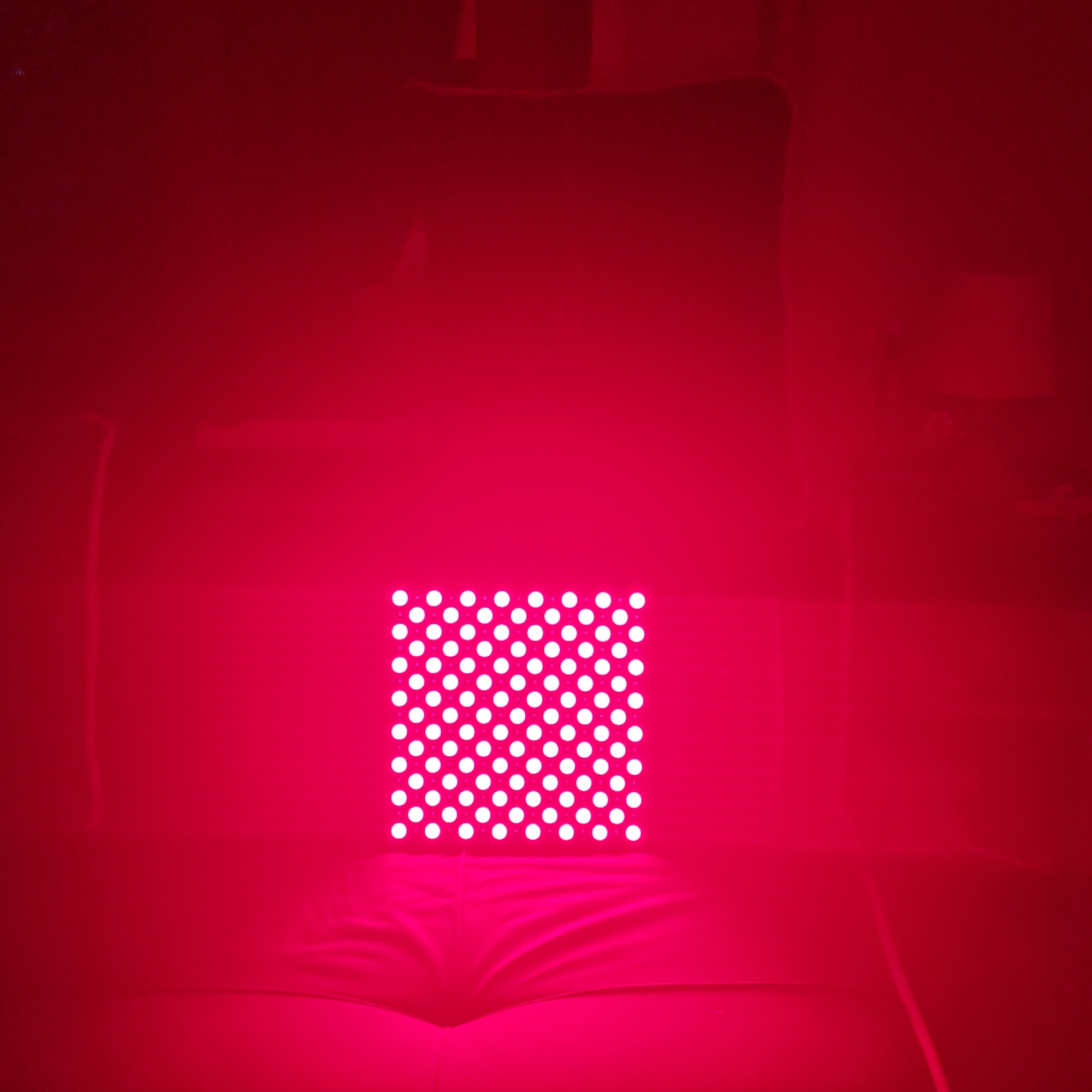 The image shows a square red light panel with an array of white circular LEDs. The panel is positioned in the center of the frame, emitting a strong red light that dominates the scene. The panel rests on a dark, possibly upholstered surface, which may be part of a couch or chair. The background is obscured by the red light but appears to be a dimly lit room with vague outlines of furniture. There are no people visible in the image. There is no information about the location being in Rostov, Russia.
