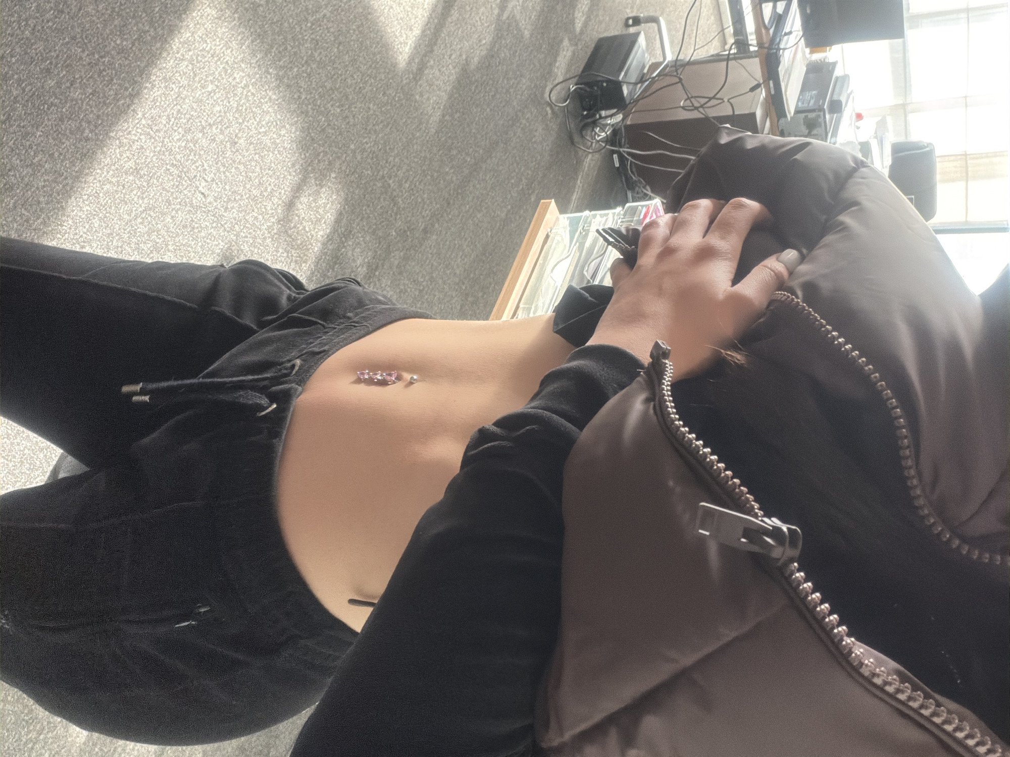 A person is seated indoors, wearing black sweatpants and a black, long-sleeved shirt partially covered by a brown puffer jacket. The person's bare abdomen is visible, featuring a navel piercing. A hand is raised towards the face. In the background, there is a cluttered area with electronics and what appears to be office equipment near a window providing daylight. A gray carpet is seen to the left. The location is unidentified, and Sofia, Bulgaria is not explicitly indicated in the image.