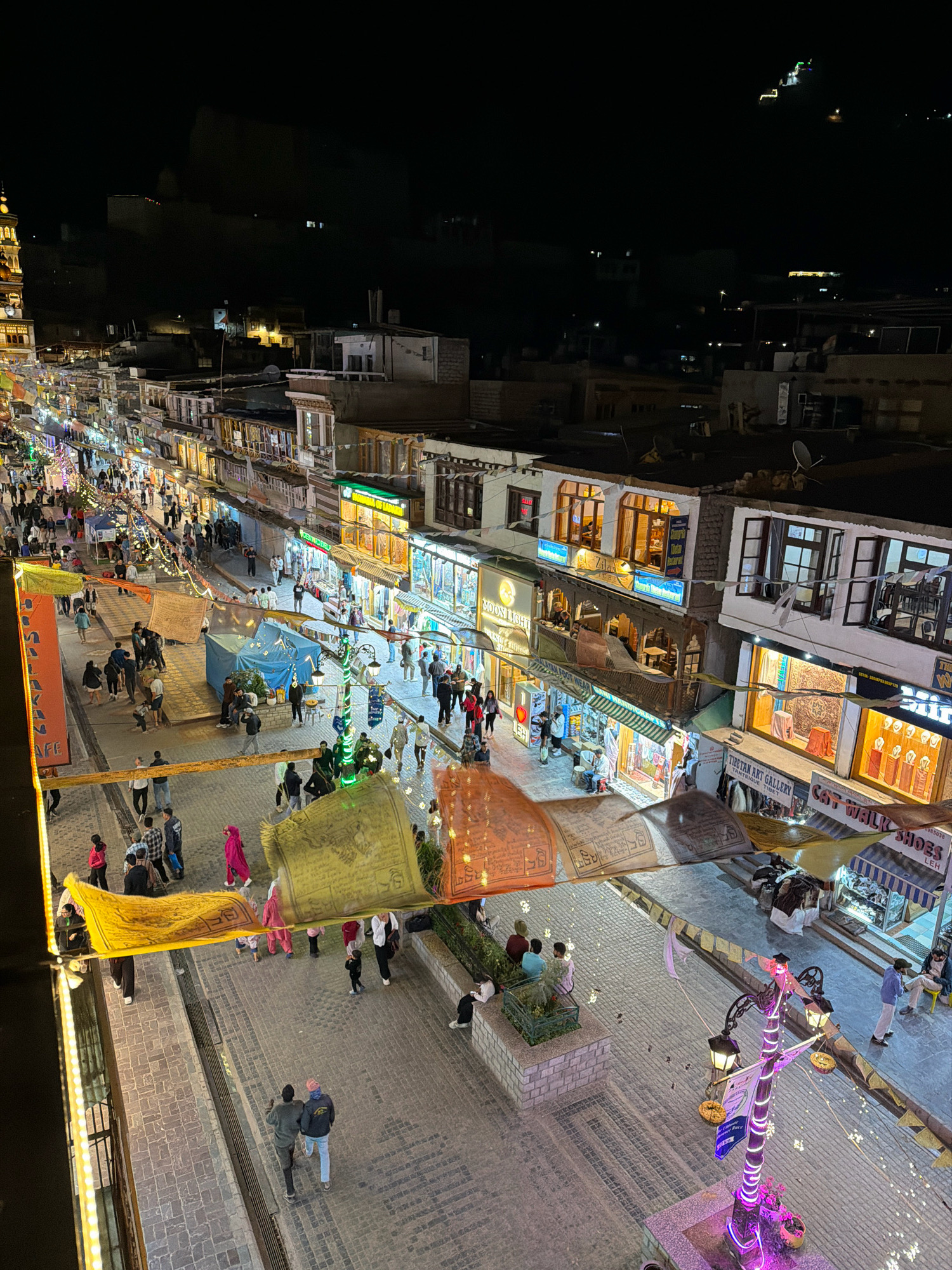 This is an elevated, wide-angle, night-time photograph of a bustling street in Leh, India. The street is lined with multi-story buildings housing shops and businesses, many of which are illuminated with warm, inviting lights. Prayer flags are strung across the street at various intervals, adding a vibrant splash of color to the scene. The cobblestone street is populated with numerous people, some walking individually, others in pairs or small groups. Several shops have displays of goods visible through their open doorways, and some have signs indicating their offerings, such as "Tibetan Art Gallery" and "Cat Walk Shoes". The overall atmosphere is lively and active, suggesting a popular commercial area. In the background, silhouetted buildings and lights on a distant hillside are faintly visible against the dark night sky.