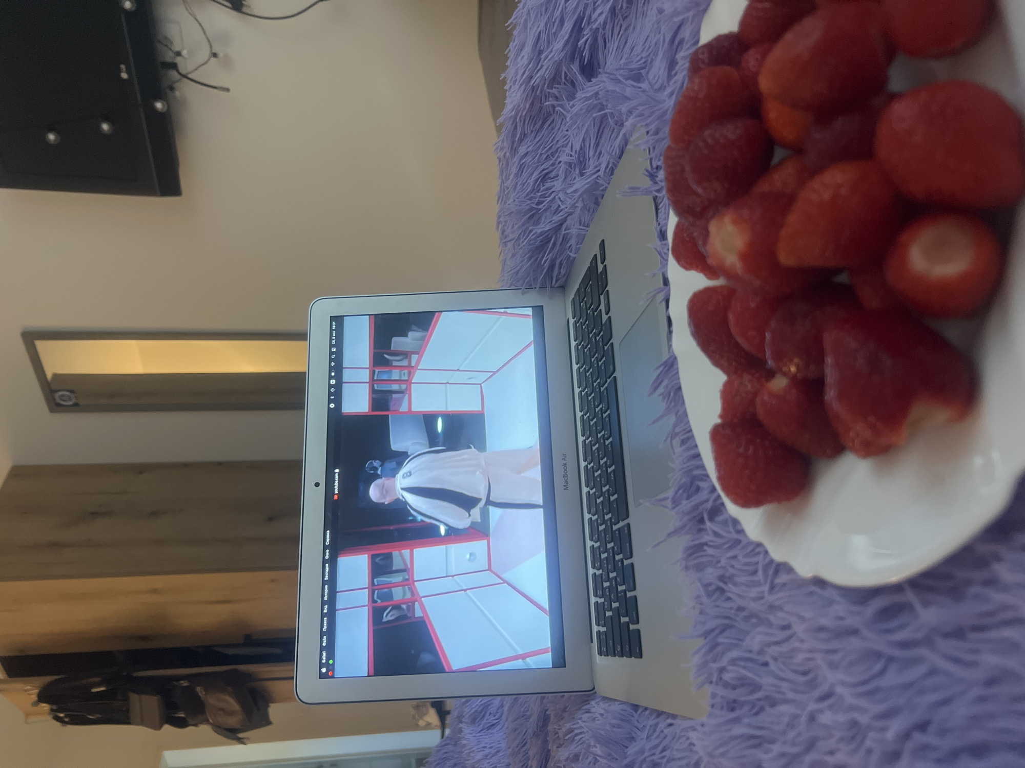The image depicts a laptop computer resting on a purple, plush surface, presumably a blanket or cushion. The laptop screen displays a video, possibly a show, featuring a man in a white and black outfit in what appears to be a staged or artificial environment. To the right of the laptop is a white plate filled with strawberries. The scene appears to be indoors, possibly in a bedroom or living room. In the background, a section of a wall and a television are visible. A light-colored wooden shelf or cabinet can also be observed with bags hanging from it. I can not determine the location in the city of Budva, Montenegro, from the provided image.