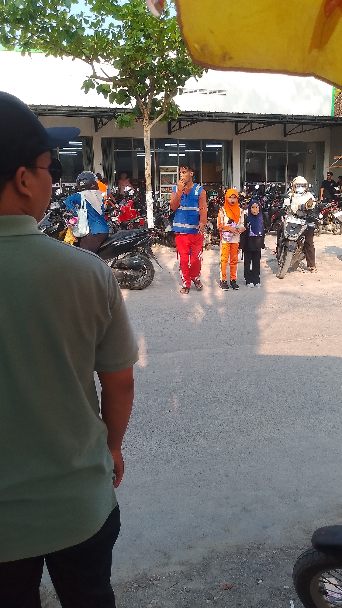 A group of people are gathered in an outdoor area, likely a parking lot, in front of a building with an awning. In the foreground, a person wearing a green shirt and black pants is facing away from the camera. Behind them, several motorcycles are parked. In the middle ground, a man in a blue vest and red pants stands and appears to be talking. Two young girls, one in an orange hijab and outfit, and another in a blue hijab and dark outfit, stand next to him. Another person wearing a white hijab and jacket is on a motorcycle to the right. The background shows the facade of the building with open doorways and awnings. The ground is paved with asphalt. The scene appears to be located in Karanglangu, Indonesia, suggested by the building's signage, though specific identifying marks are not clearly visible in this crop.