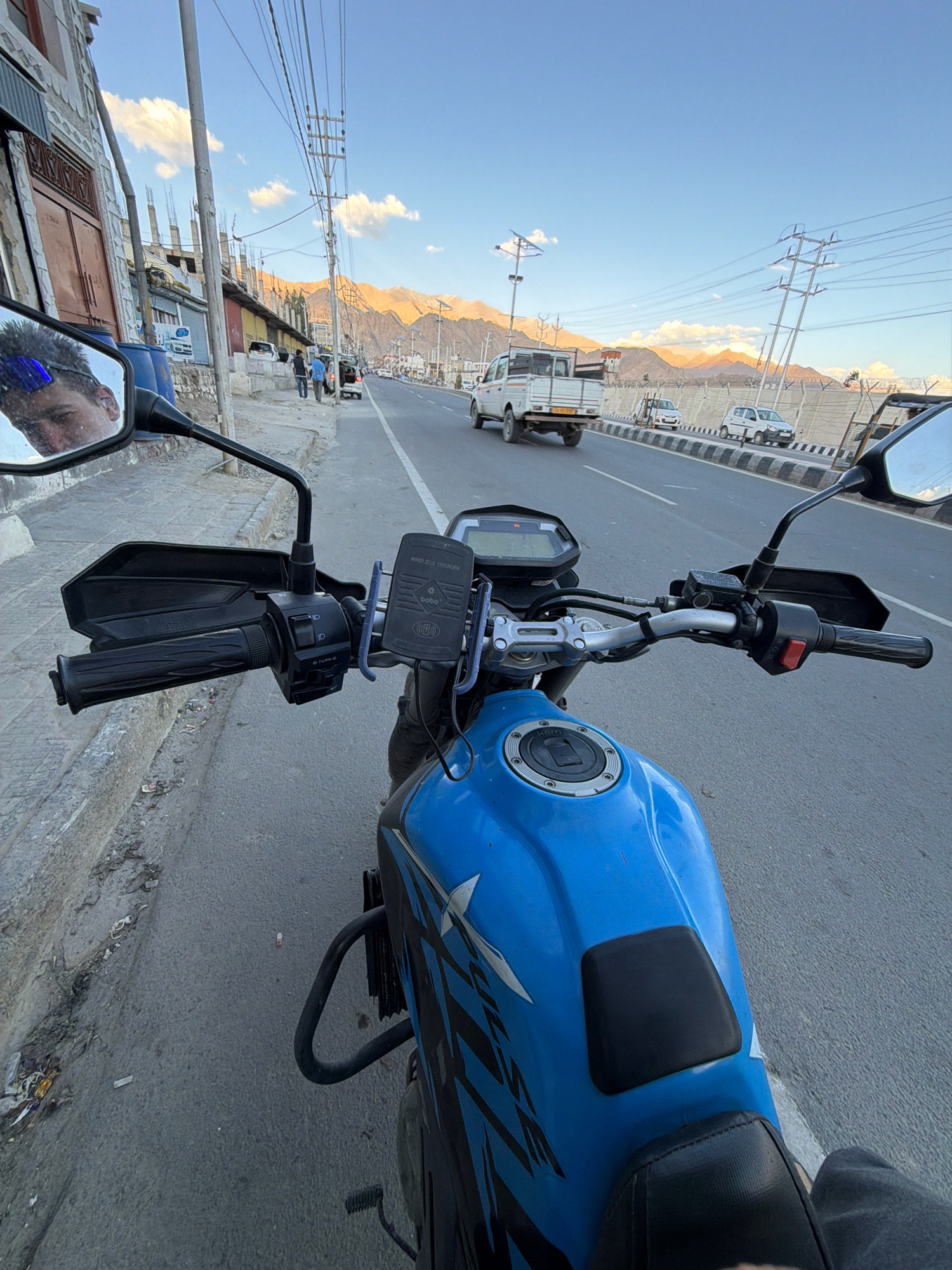 The image is taken from the perspective of someone riding a blue motorcycle on a road in Leh, India. In the left mirror, a person's face is reflected. The motorcycle has black handguards and a wireless charger mounted on the handlebars. The road is paved and has a sidewalk on the left. Buildings line the left side of the road, some appearing under construction. In the background, mountains are visible with warm light shining on them. Several vehicles, including a white pickup truck and white cars, are on the road. Overhead power lines are also visible. The sky is blue with scattered clouds. The scene appears to be at dusk or dawn.