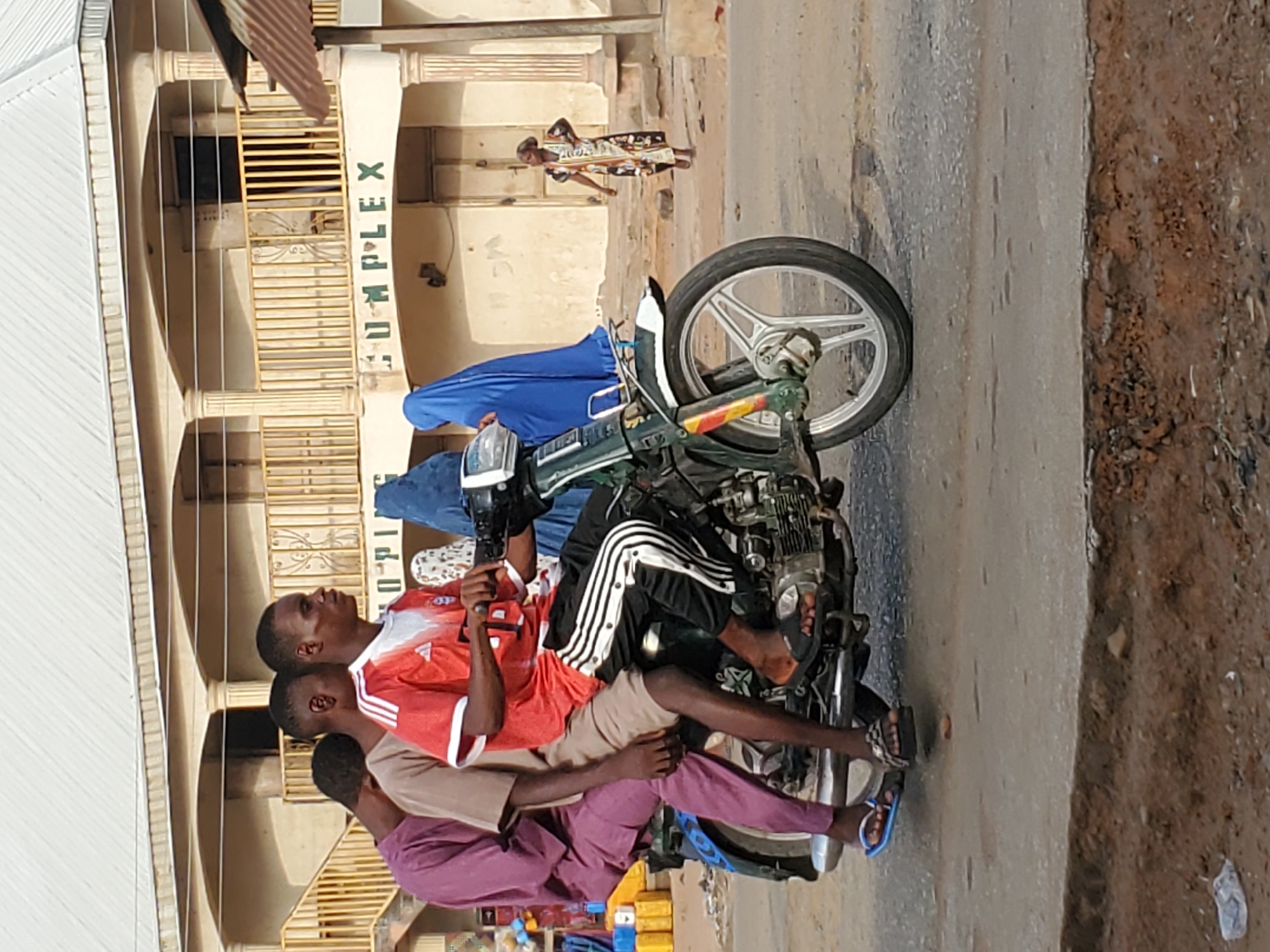 In an urban street scene, a green motorcycle is carrying at least four individuals. The person at the front, likely the driver or main passenger, wears a red and white striped t-shirt. Behind them, another person wears a light brown shirt, and a third, at the very back, is dressed in a purple garment. A fourth individual, obscured by the others and the motorcycle, is visible in a blue garment. They are either riding or stopped on an asphalt road next to a dirt shoulder.
In the background, a multi-story building with a light-colored facade features balconies with yellow grilles and pillars. The word "COMPLEX" is written vertically in green letters on the side of this building. Further down the street, a child in a patterned dress walks on a sidewalk. The scene is brightly lit by daylight, suggesting clear weather. The overall setting, including the architecture and the common practice of multiple passengers on a single motorcycle, is consistent with a location in Mararaba, Nigeria. To the bottom left, some yellow containers are partially visible, possibly indicating a street vendor or shop.