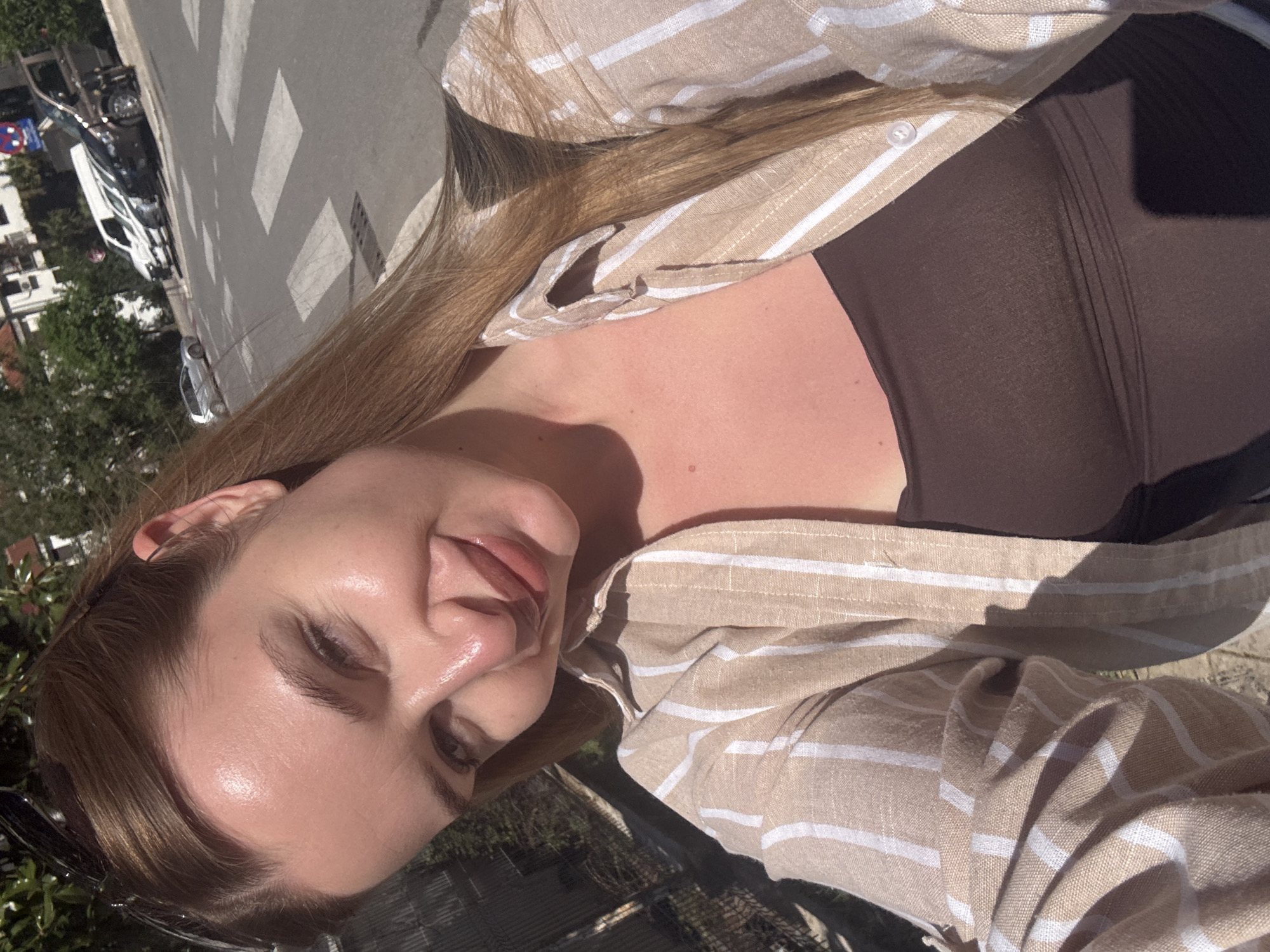 A close-up selfie, rotated counter-clockwise, features a young woman outdoors on a sunny day in what appears to be Budva, Montenegro. She has light brown hair and fair skin, captured looking towards the camera with a gentle smile. She wears a beige or tan striped long-sleeved button-up shirt with white horizontal stripes, layered over a dark brown or black top. Sunglasses are visible resting on her head.
The bright sunlight casts distinct shadows, suggesting it's midday or early afternoon. The background reveals an urban street scene with an asphalt road featuring a white pedestrian crossing. Parked vehicles, including a white van, another car, and a motorcycle, are visible. Green trees and building facades line the street. A blue circular traffic sign with a white arrow is visible on the left side of the background. The woman's chest and neck show a slight flush, possibly from sun exposure. No discernible text is visible other than the arrow on the traffic sign.