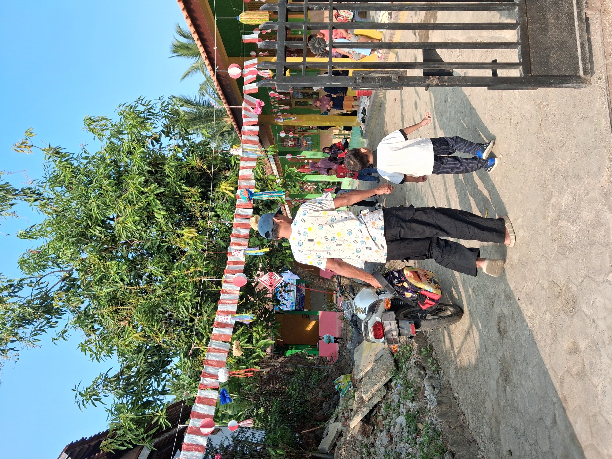 In Bantarkawung, Indonesia, a man and a young boy walk together on a paved path towards a school building. The man, from behind, wears a patterned t-shirt, black pants, and a blue cap. He carries a backpack with a colorful design on his left shoulder. The boy, also viewed from behind, wears a white t-shirt with black trim and striped pants. They are walking next to a white motorcycle parked on the left side of the path. The environment features a building with a green facade decorated with festive red and white streamers and various hanging ornaments. A black metal gate is positioned on the right side of the path. In the background, other people, including adults and children, are visible near the school entrance. The scene is set under a clear blue sky with lush green trees and palm trees providing a natural backdrop. Debris, including concrete blocks and rocks, is scattered on the left side of the path, suggesting ongoing construction or renovation.