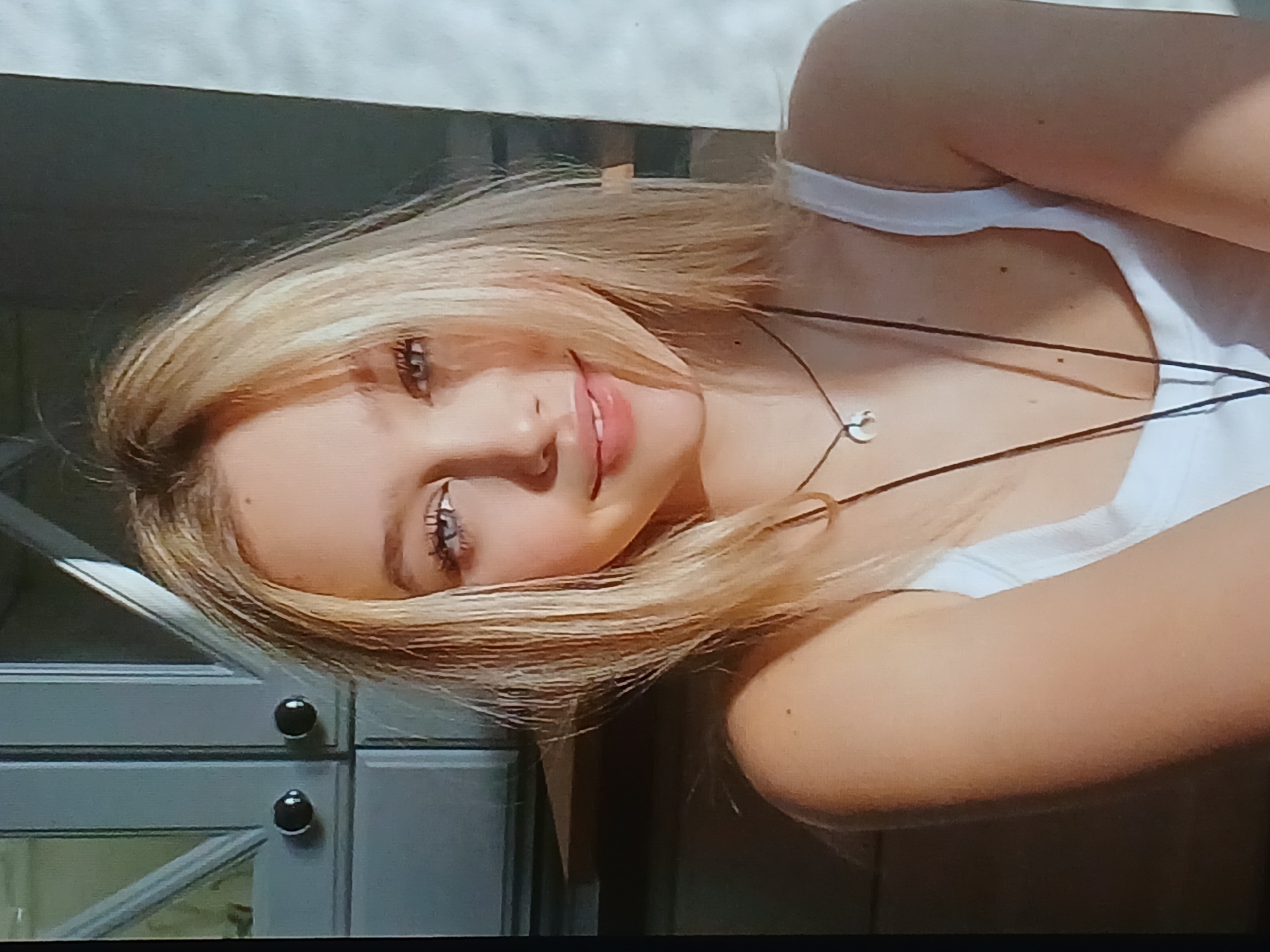 A blonde young woman with blue eyes is looking directly at the camera with a slight smile. She is wearing a white tank top and a necklace with a crescent moon pendant. Her hair is shoulder-length and slightly wavy. The background appears to be an indoor setting, possibly a room with light-colored furniture and a window that is casting a shadow. There are no discernible elements in the image that would indicate a specific location within the city of Dnipro, Ukraine.