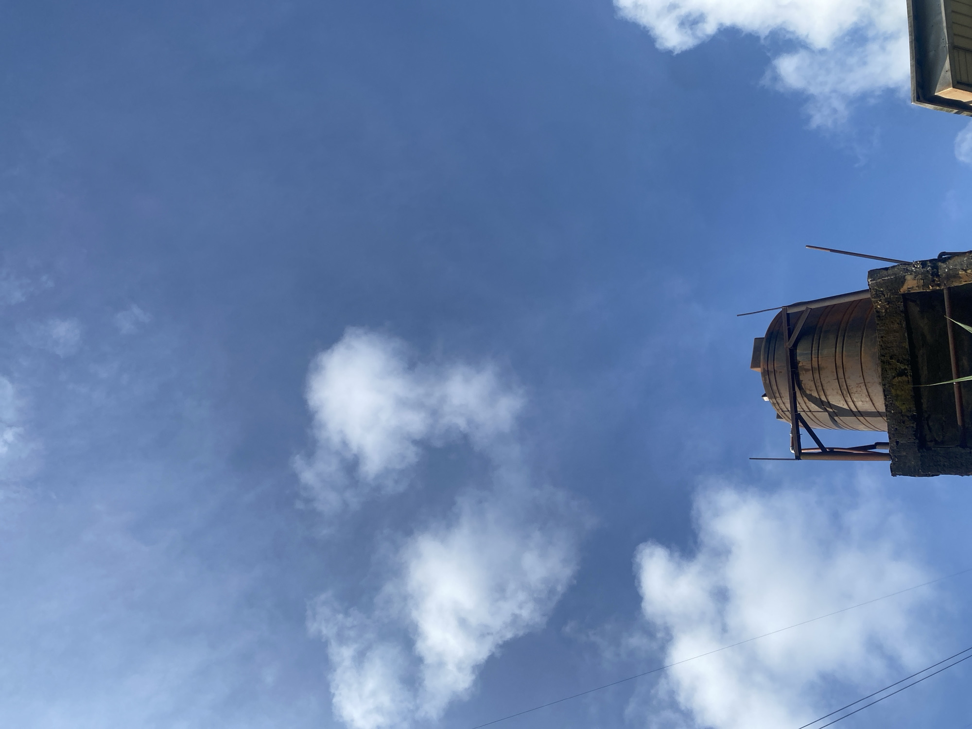 The image shows a water tank on top of a concrete structure against a bright blue sky with scattered white clouds. The water tank appears to be made of metal and has a rusty, weathered surface. It is supported by a metal frame. There are also some thin wires running diagonally across the sky. The scene is likely captured from a low angle, looking upwards. The overall impression is one of an urban or semi-urban environment in Ado, Nigeria, where such water storage systems are common.