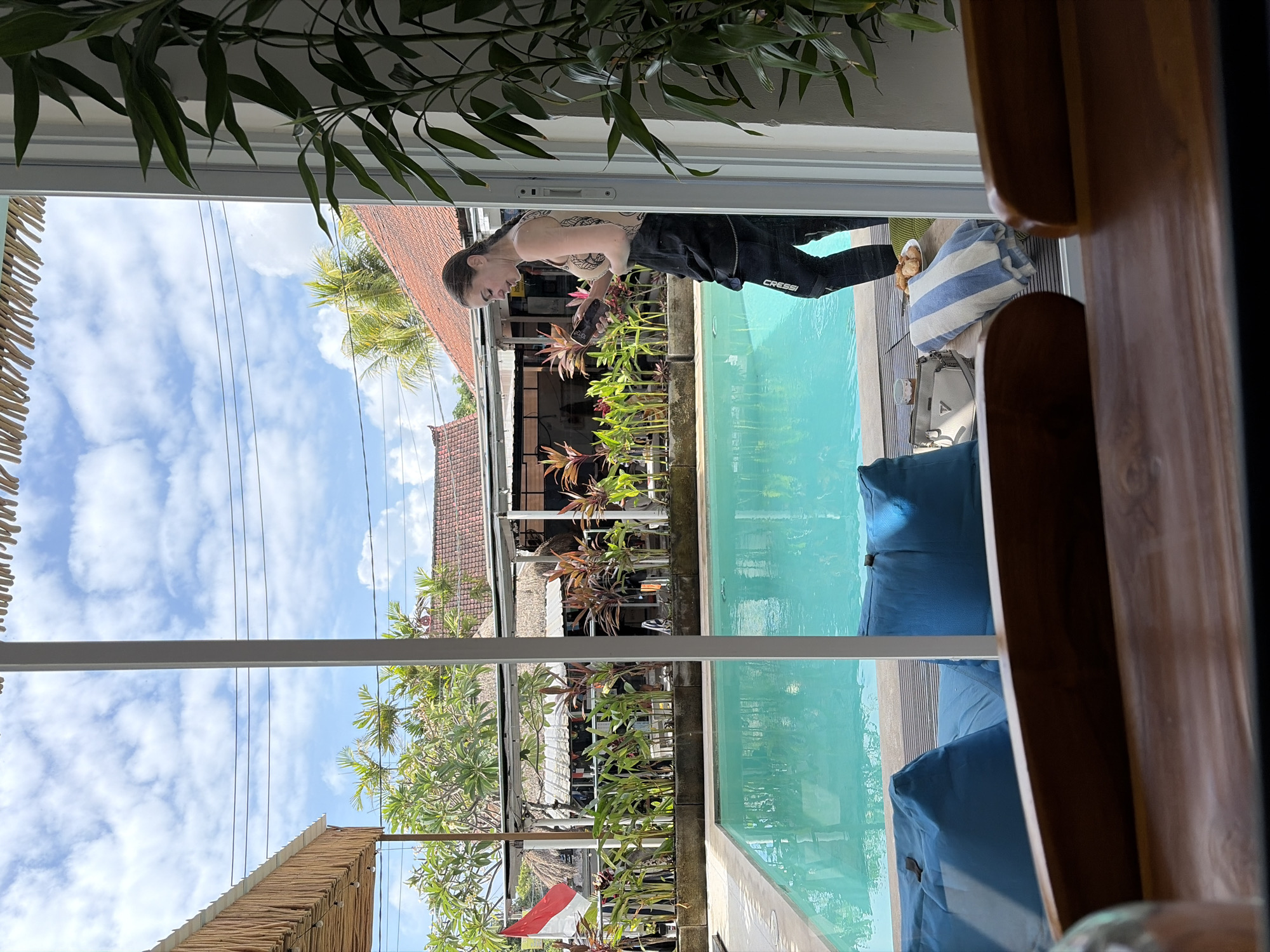The image depicts an outdoor scene, likely a resort or villa, with a swimming pool as the central focus. A woman, standing near the pool, is looking at her phone. She is wearing a patterned top and dark pants. Beside her, on a table, are a white handbag, a folded towel with blue stripes, and a plate of food. The swimming pool is turquoise blue, surrounded by a deck and vegetation. In the background, there are buildings with terracotta roofs, a red and white flag, and palm trees, suggesting a tropical location. The overall atmosphere appears relaxed and idyllic, possibly in Kubu, Indonesia, given the architectural style and lush greenery. The foreground shows a blurred wooden surface, likely a table, suggesting the image was taken from inside a building looking out.