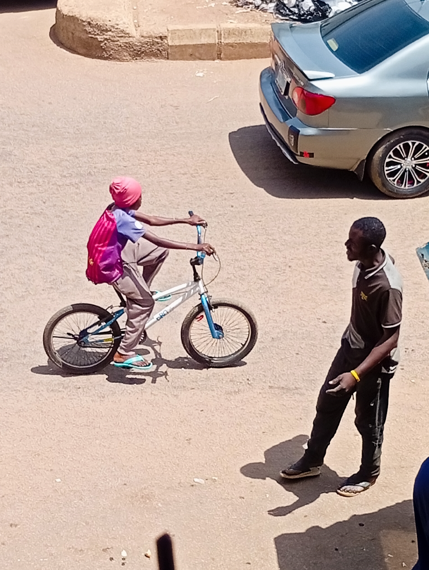 A person wearing a pink headscarf and a pink and purple backpack rides a bicycle on a dirt road in Bukuru, Nigeria. The bicycle is white and blue with black tires. A second person, a man wearing a dark brown and gray shirt and black pants, stands on the road to the right of the cyclist, facing away from the camera. A silver car is parked on the right side of the image. The ground is a light brown dirt surface, and there is a raised, tan-colored concrete structure in the background. The lighting indicates a sunny day, casting distinct shadows.