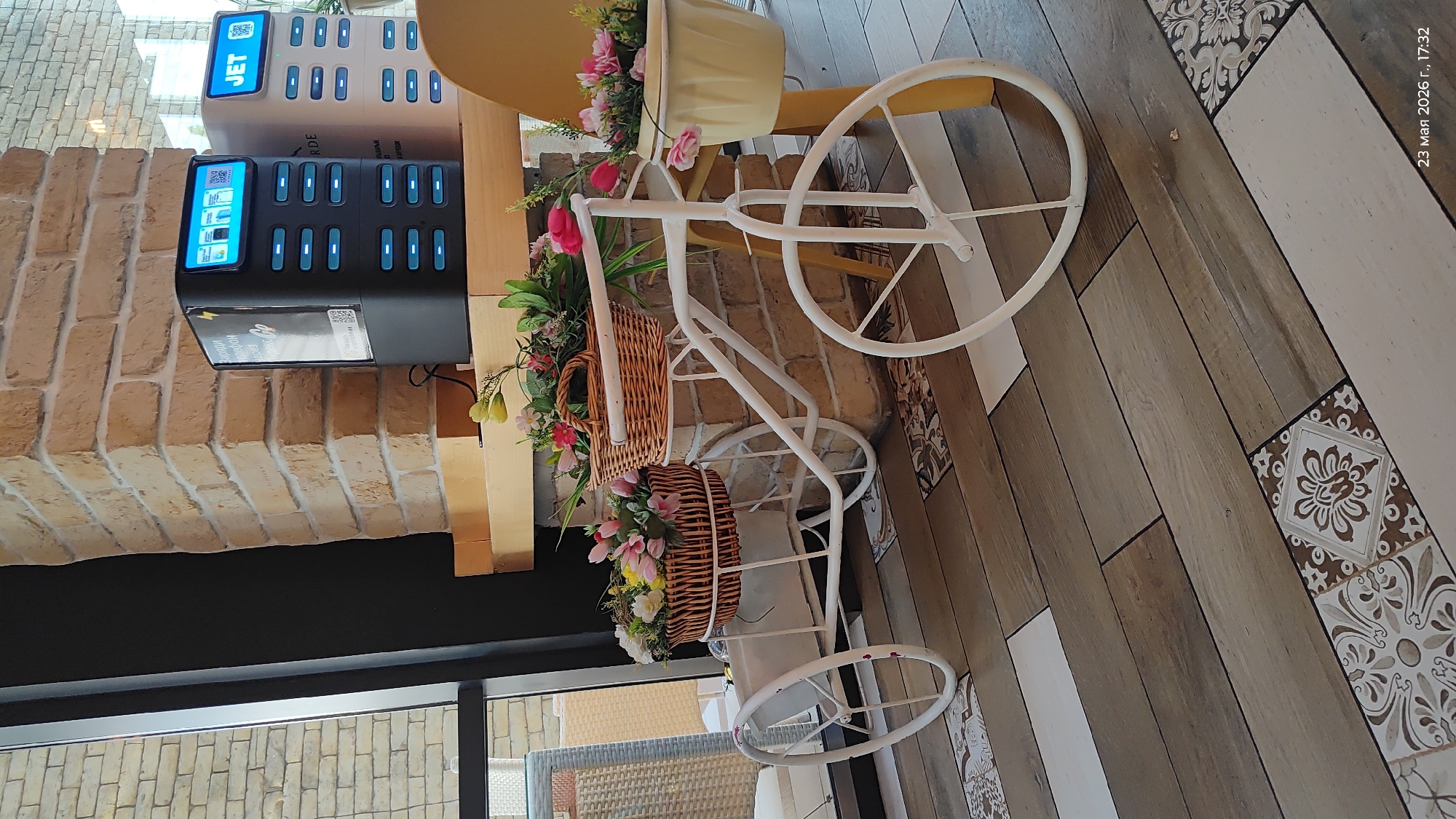 The image shows a decorative white bicycle with baskets filled with flowers, placed indoors, likely in a cafe or restaurant. A charging station with "JET" branding is attached to a brick wall, suggesting a service provided by the establishment. The flooring is a mix of wood-look planks and decorative tiles. The date stamp in the upper right corner indicates the image was taken on March 23, 2026. The presence of the "JET" charging station suggests this might be Almaty, Kazakhstan, where the service is known to operate.