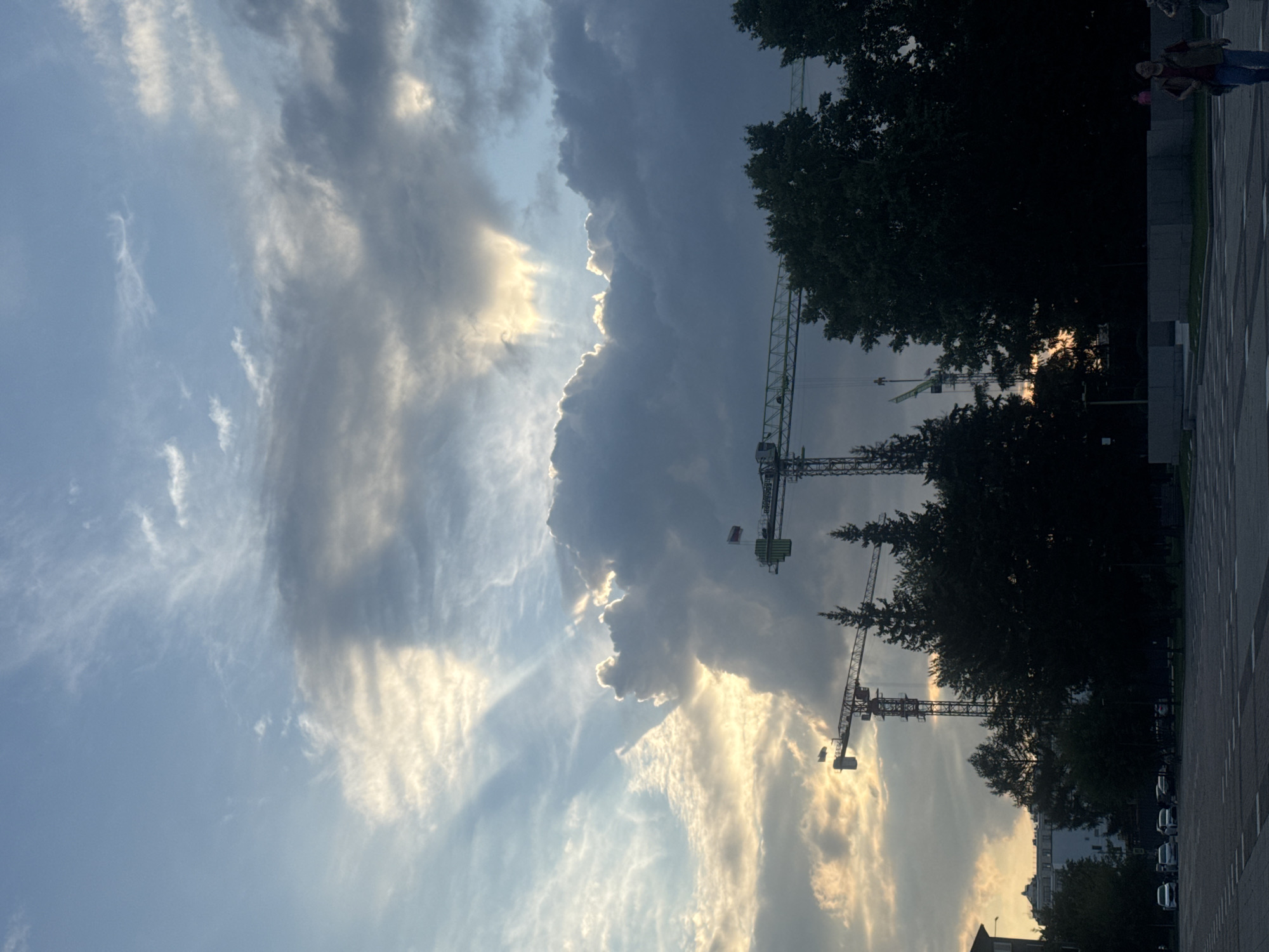 This is a vertical, eye-level, medium-distance shot of a construction site under a dramatic sky, likely in Irkutsk, Russia, given the presence of construction cranes and a park-like area. In the foreground, there is a paved area with white markings, resembling a parking lot or plaza. Beyond this, silhouetted dark green trees form a dense line across the middle ground. Two large construction cranes dominate the upper middle ground, one taller and red, the other shorter and lighter colored. The red crane has a flag with horizontal red and white stripes at its top. The sky is a dynamic mix of bright sunlight breaking through dark, heavy clouds, creating rays of light. Some wispy white clouds are also present. In the far background, buildings are visible, and a few cars are parked along a street to the left. Two individuals are walking in the lower right foreground, one wearing a vest over a dark shirt and jeans, the other partially visible. The overall atmosphere is one of an active urban environment juxtaposed with a powerful natural sky.