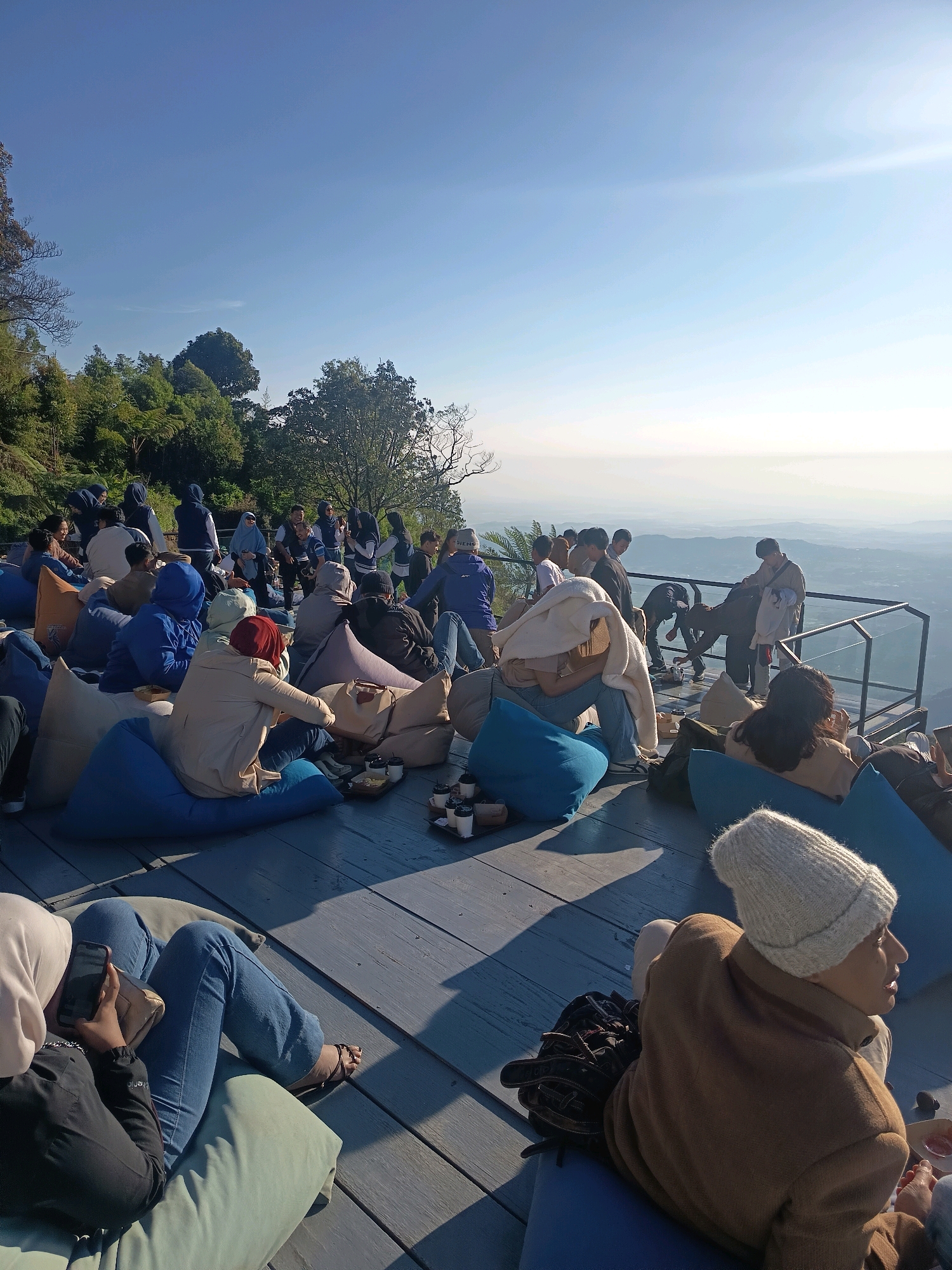 In Keditan, Indonesia, a group of approximately 30 people are gathered on an elevated wooden deck overlooking a misty valley. The scene appears to be a cafe or viewing area, with many individuals seated on large beanbag chairs. The atmosphere is relaxed, with people dressed casually in light clothing, suggesting a warm climate. Some are holding phones, engaging in conversation, or looking out at the view. The deck is bordered by a metal railing, indicating a significant drop. Lush green trees and foliage surround the area, and the sky is a clear, bright blue, transitioning to a hazy horizon. The overall impression is one of a tranquil outdoor gathering enjoying a scenic vista.