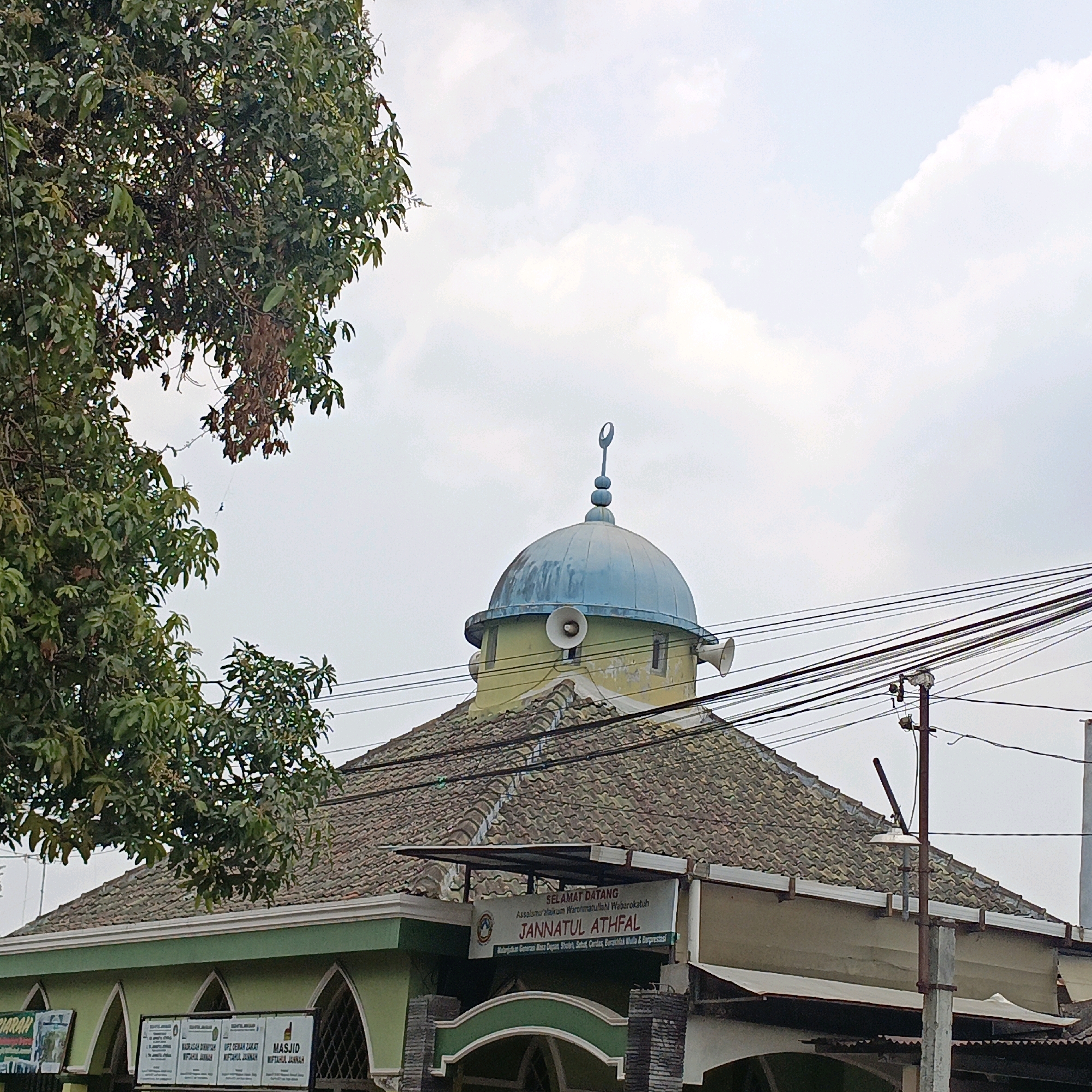 The image shows the exterior of a mosque, identified by a sign as "JANNATUL ATHFAL", located in Salatiga, Indonesia. The mosque features a light blue dome topped with a crescent moon and star symbol. Two loudspeakers are mounted on the sides of the dome's base. The roof of the mosque is covered with brown tiles. A prominent tree with green leaves is visible in the upper left portion of the image, partially obscuring the sky. Electrical wires run across the scene, supported by a utility pole. Signs with text in Indonesian are visible on the lower part of the building. The sky is overcast with clouds.