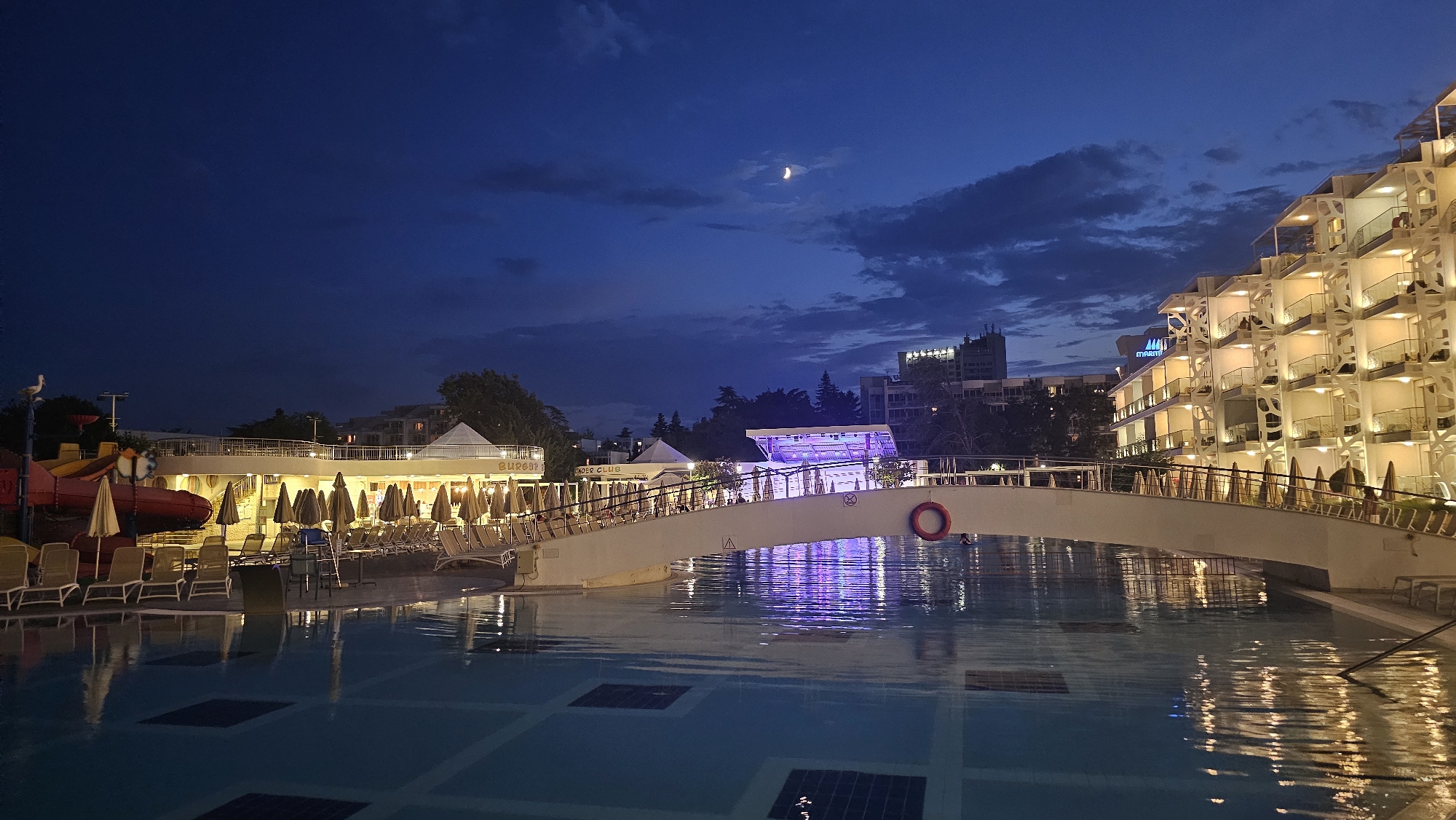 The image depicts a resort scene at dusk in Obrochishte, Bulgaria, with a crescent moon visible in the deep blue, cloudy sky. In the foreground, a swimming pool reflects the ambient light. A white pedestrian bridge spans the pool, illuminated by purple lights from a stage or event area underneath. To the left of the bridge, a white building with numerous open umbrellas and lounge chairs suggests a dining or relaxation area. A children's water slide is also visible. To the right of the bridge, a modern, multi-story hotel building glows with interior lights, its balconies and white facade prominently featured. The overall atmosphere is calm and serene, with the resort appearing quiet as night settles in.