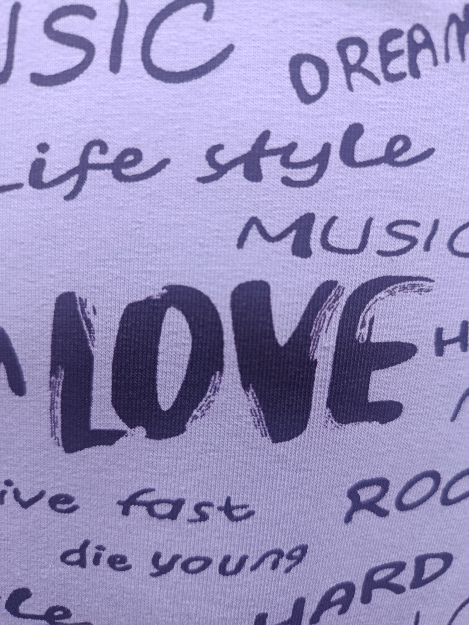 The image is a close-up, high-angle, detailed shot of a light purple fabric with dark purple lettering. The fabric appears to be a t-shirt or similar garment. The lettering consists of various words, many of which are short phrases. Prominently displayed in large, bold font is the word "LOVE". Other visible words include "MUSIC", "DREAM", "life style", "ive fast", "die young", "ROC", and "HARD". The lettering is in a mix of fonts, with some words in a more stylized, script-like font and others in a blocky, uppercase font. The texture of the fabric is visible, showing the knitted pattern. There are no people or the environment of Sertolovo, Russia are visible in this image.