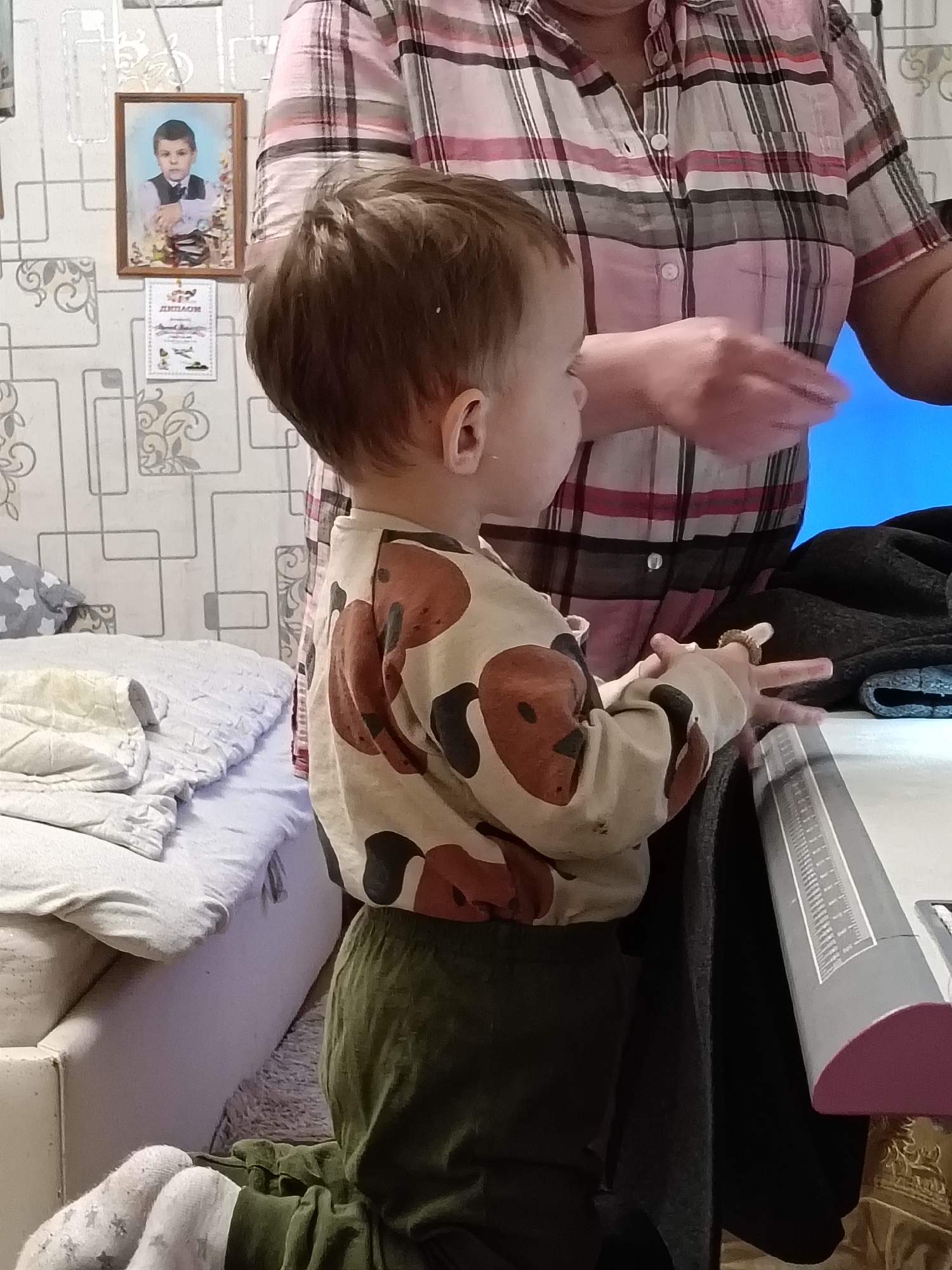 A child stands next to a table in a room in Красный Маяк, Russia. The child wears a beige shirt with brown patterns and dark green pants. An adult wearing a pink and gray plaid shirt stands to the right of the child. A bed is visible to the left of the child. A picture frame hangs on the wall, and there is also a certificate displayed below it.