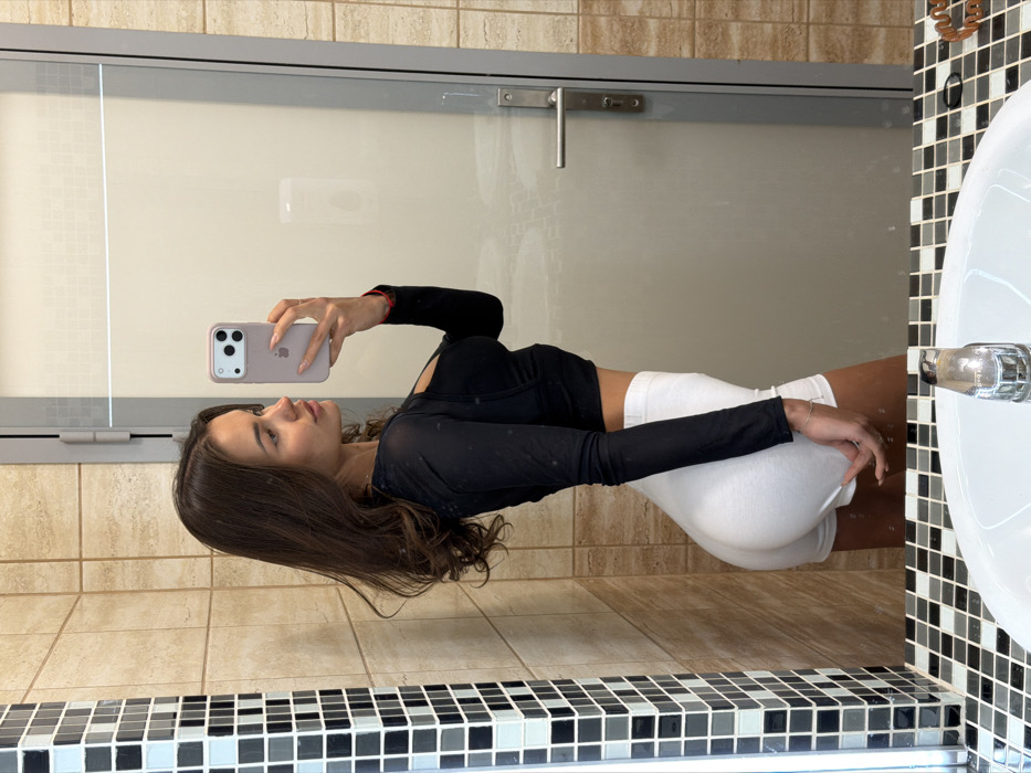 A young woman poses in a bathroom mirror, holding up a smartphone to capture her reflection. She is wearing a black long-sleeved top and tight white shorts. Her long, dark hair cascades over her shoulders. The bathroom has beige wall tiles and a mosaic-tiled countertop. A glass-paneled door with a silver handle is behind her. A white sink is visible on the right side of the frame. The overall image has a slightly rotated perspective.