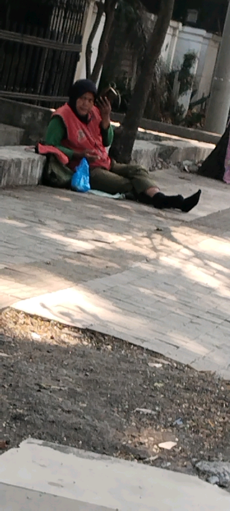 In Semarang, Indonesia, a person wearing a hijab and a pink vest over a green shirt sits on a concrete ledge. They hold a dark object in their right hand and appear to be looking at it. A blue plastic bag lies on the ground in front of them. The person is wearing dark pants and black socks. The background shows trees, a fence, and part of a building. The ground is paved with concrete and gravel, with shadows cast by trees.