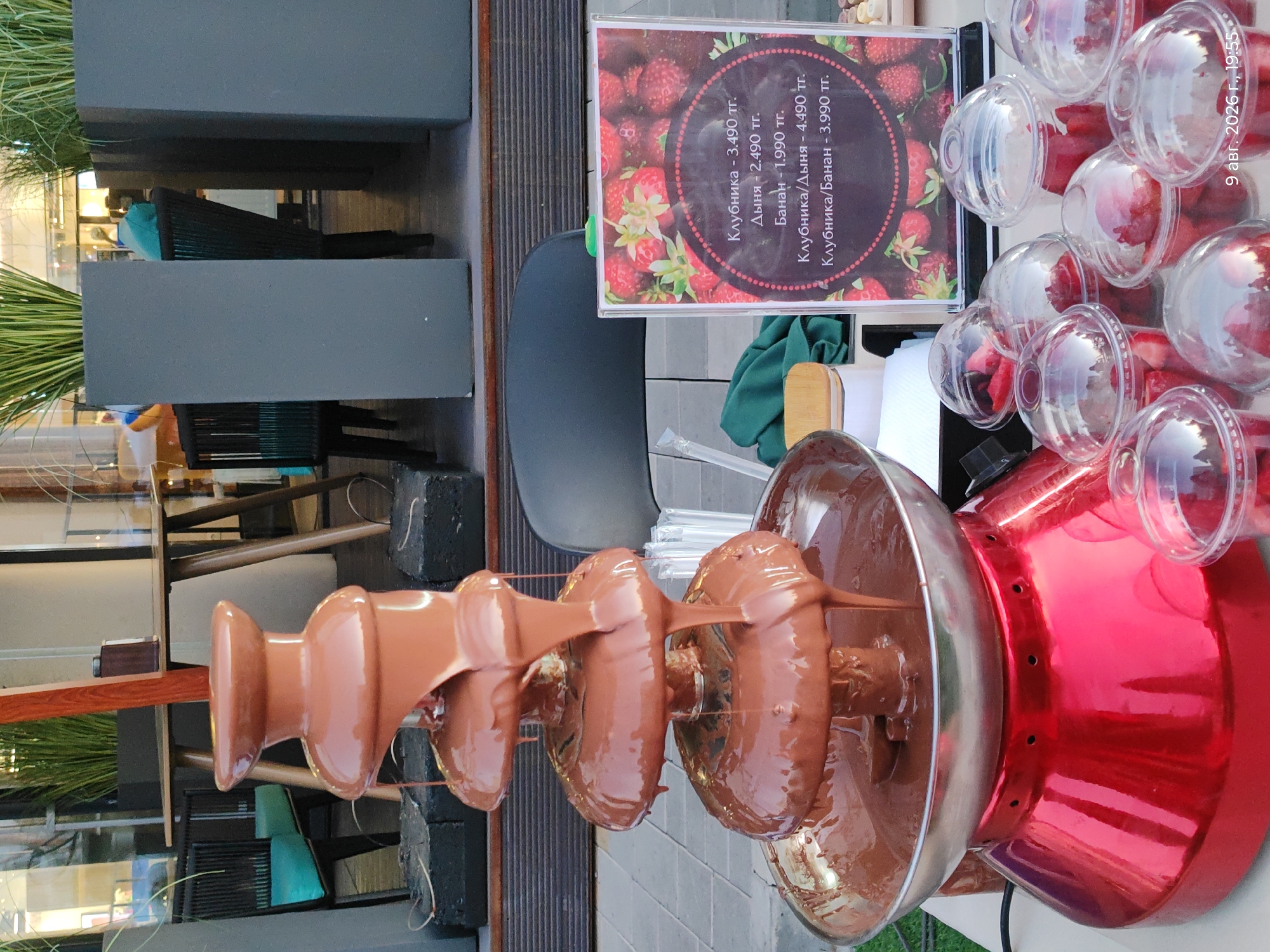 The image depicts a chocolate fountain with melted chocolate cascading down its tiers. In the foreground, several clear plastic cups filled with strawberries are arranged on a table. To the right, a sign displays prices for strawberries, melons, bananas, and combinations thereof, in what appears to be Russian currency. The background shows an outdoor seating area with tables, chairs, and large planters containing greenery. The overall setting suggests a cafe or outdoor market. The date and time stamp at the bottom right corner indicates "9 авг. 2026 г., 19:55", which translates to August 9, 2026, at 7:55 PM. The city is identified as Astana, Kazakhstan.