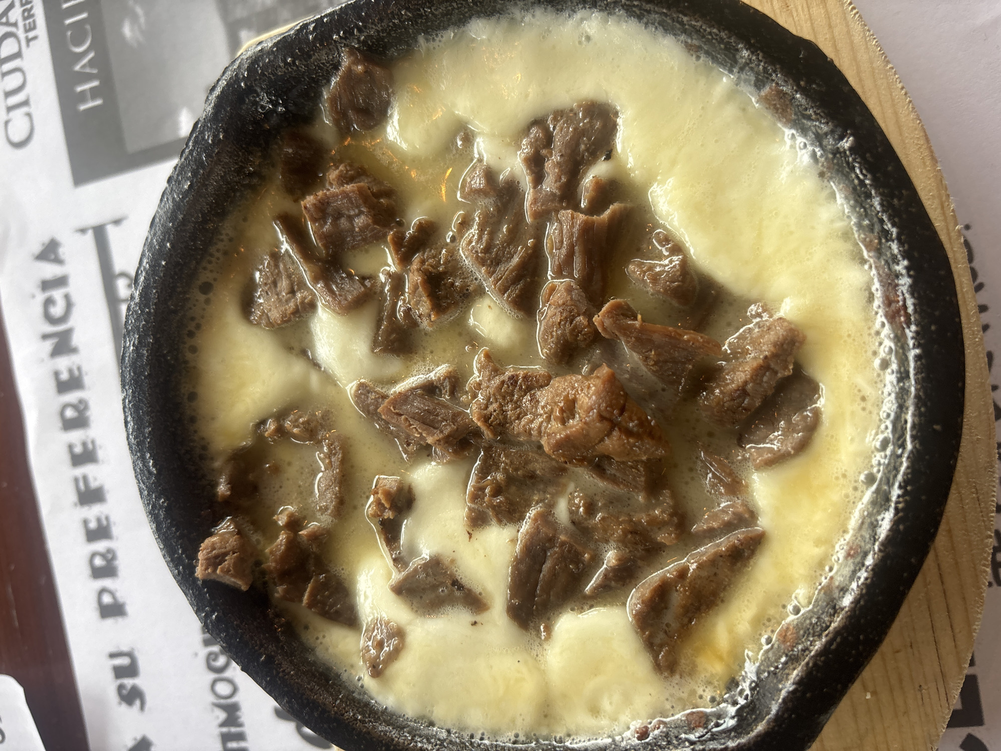 A top-down, medium shot shows a hot and bubbly dish of beef and cheese, likely served in Puebla, Mexico. The dish is presented in a small, dark-colored, possibly cast-iron skillet-type vessel. The cheese is pale yellow, melted, and bubbly. Chunks of cooked, seasoned beef are scattered throughout the cheese. The skillet is sitting on a light-colored wooden coaster or board. Part of a paper placemat or menu is visible, displaying the word "CIUDAD" and the phrase "SU PREFERENCIA" in Spanish. The setting appears to be a restaurant in Puebla, Mexico. The atmosphere suggests a casual dining experience.