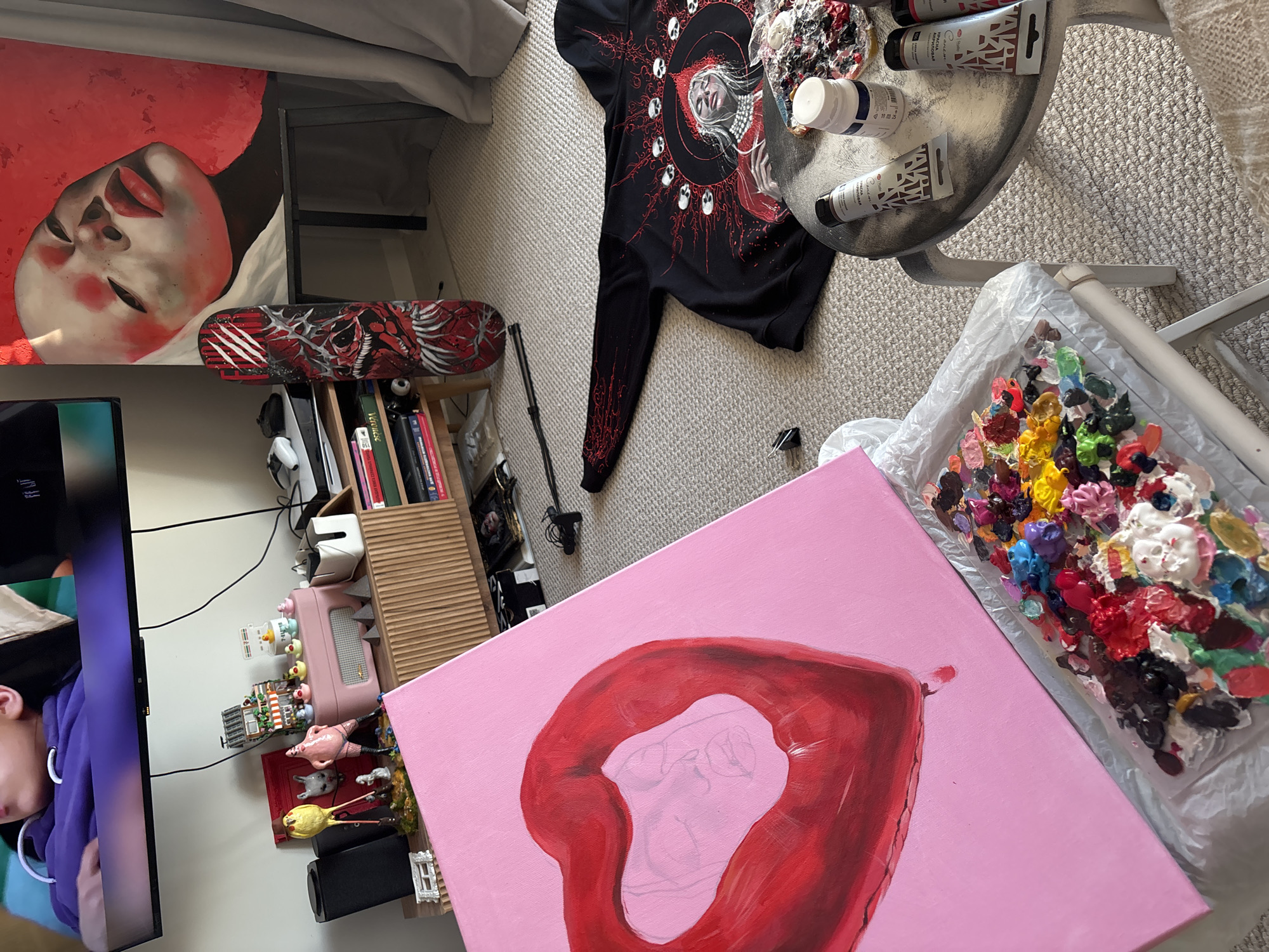 This media file captures an indoor scene, likely a personal art studio or creative space in Krasnoyarsk, Russia. The environment is rich with artistic elements and personal decor.
The central activity appears to be painting. In the foreground, a pink canvas displays a partially painted red, heart-shaped object, possibly a balloon or lips, with a faint pencil sketch visible inside. Adjacent to this, a clear tray or palette holds numerous blobs of vibrant acrylic paint in various colors, including red, yellow, blue, green, white, black, purple, and brown. On a small, round, metallic-looking table, several tubes and jars of paint are visible, with brands like "SPACIA ACRYLIC" and "MASTERS ACRYLIC" legible on their labels.
Other notable objects include a large painting leaning against furniture, depicting a woman's face with striking red lips against a red background. A black long-sleeved shirt or sweatshirt with an intricate red and white graphic design, featuring a central figure surrounded by skulls, is spread on the carpet. A skateboard deck with a red and black skull-themed design rests near a bookshelf filled with books, one of which appears to have "VERONICA" on its spine.
The room also contains a television displaying a person in a purple hoodie, suggesting media consumption alongside the creative work. Various decorative items are scattered around, such as a pink ribbed container with small figurines (including duck-like characters), miniature houses, and animal figures. The floor is covered with a light-colored carpet, and the overall lighting suggests it is daytime.