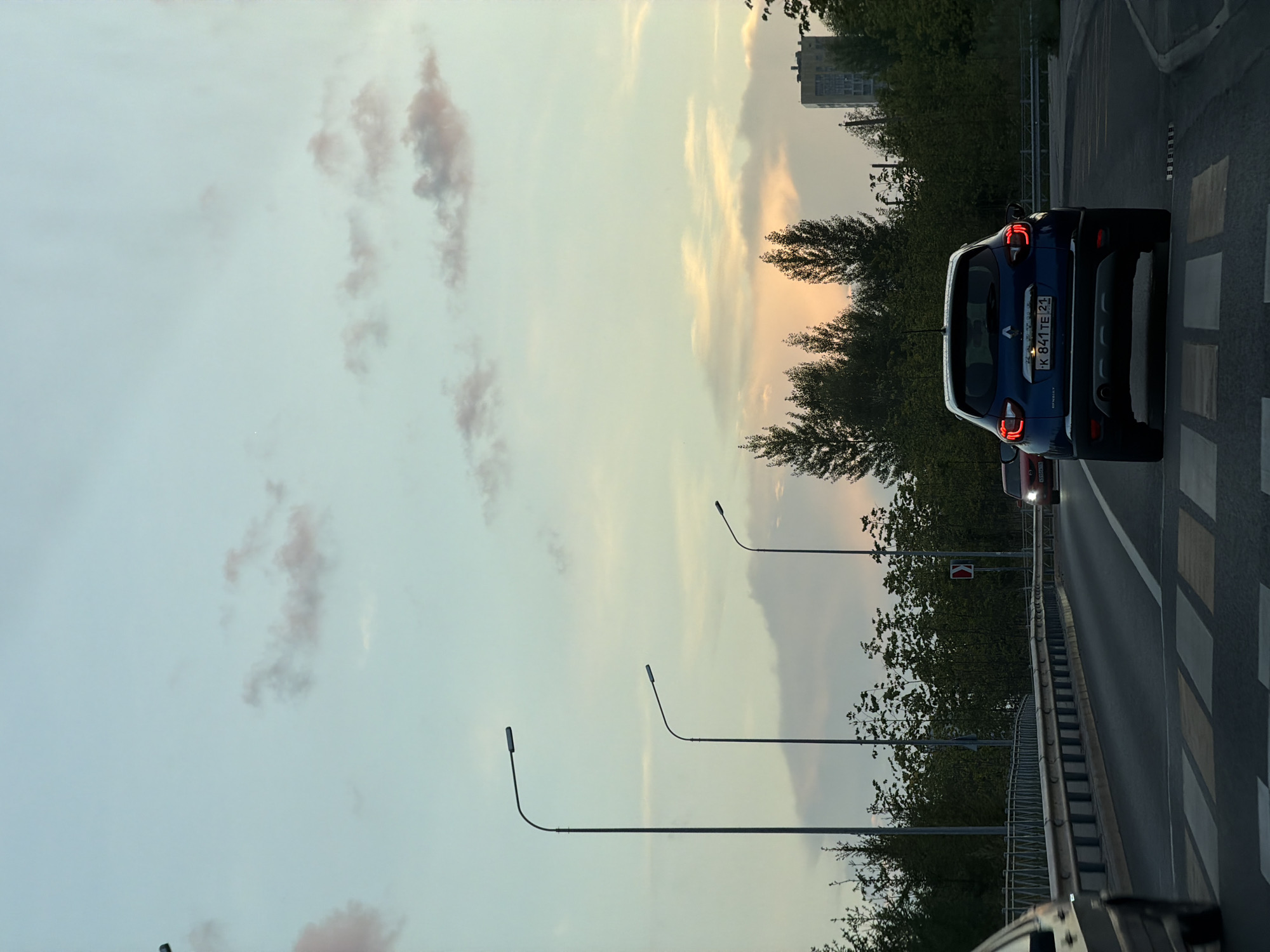 A dark blue Renault Captur SUV is stopped at a pedestrian crossing on a multi-lane road in Kazan, Russia. The car's license plate reads K 841 TE 21. The sky above is a pale blue with wispy pink clouds, suggesting either sunrise or sunset. Tall streetlights line the road, and a bridge railing is visible on the left. Trees and a tall building are in the background. A red car is visible further up the road. The scene appears to be an urban or suburban setting during twilight.