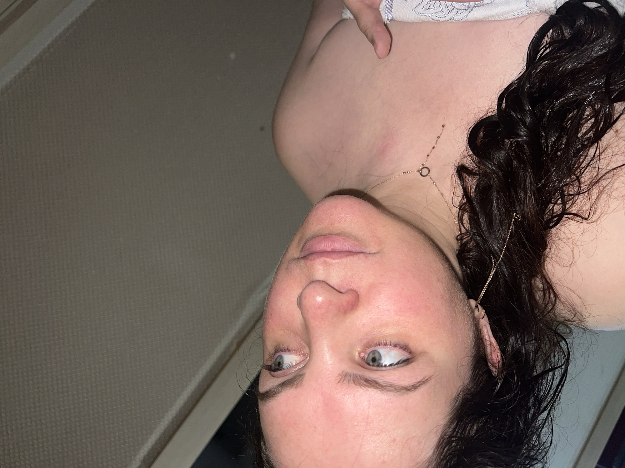 A close-up, angled portrait shows a person with light skin and dark, curly, possibly wet hair. The person is wearing a delicate gold necklace. The eyes are light-colored, and the facial expression is neutral. The background is a textured, light-colored wall. The image does not contain enough information to determine the location as Noviy, Russia.
