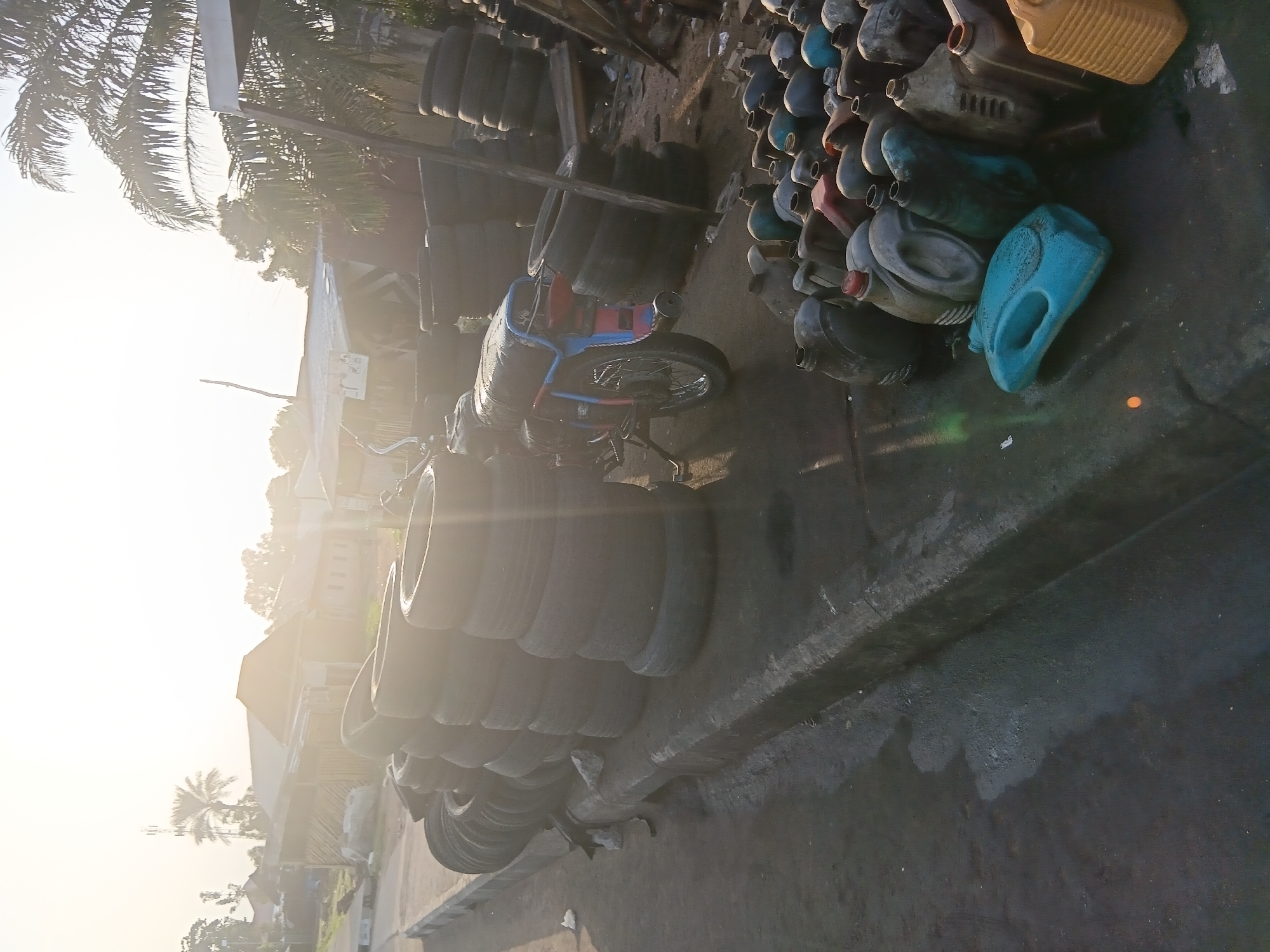 In Jalingo, Nigeria, a street scene is captured with a focus on a collection of discarded items. The foreground is dominated by a large assortment of plastic containers, primarily used for liquids, varying in size and color. These containers are densely packed and appear to be gathered for disposal or recycling. Behind them, a motorcycle is parked, its details partially obscured by the surrounding objects. The mid-ground features stacks of used tires, piled high on the side of the road. Further in the background, buildings are visible, suggesting a residential or commercial area. The environment appears somewhat unkempt, with debris and dust evident on the ground. The overall lighting suggests it is either early morning or late afternoon, with the sun low in the sky casting a bright haze.