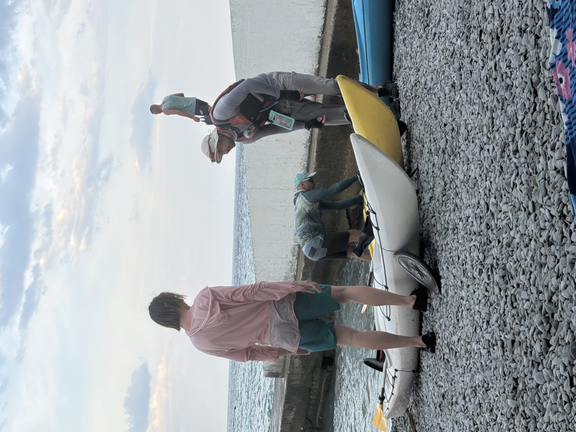 In Partenit, Ukraine, on a pebbled shoreline, a group of people prepare for kayaking. The sea, with calm waves under a cloudy sky, is visible in the background. Three kayaks, one white, one yellow, and one blue, are lined up on the shore.
A person with medium-length, dark hair, wearing a light pink hooded shirt and teal shorts, stands facing away from the camera, looking towards the water. To their right, a person wearing a hat, gray long-sleeved shirt, and life vest is bent over a kayak. Behind them, another individual in a greenish-blue shirt and dark shorts is also leaning over a kayak. Further in the background, a person wearing a light blue tank top and dark shorts stands with a bag.
The scene appears to be a preparation for a kayaking activity, with the people engaged in tasks related to launching or organizing the kayaks.