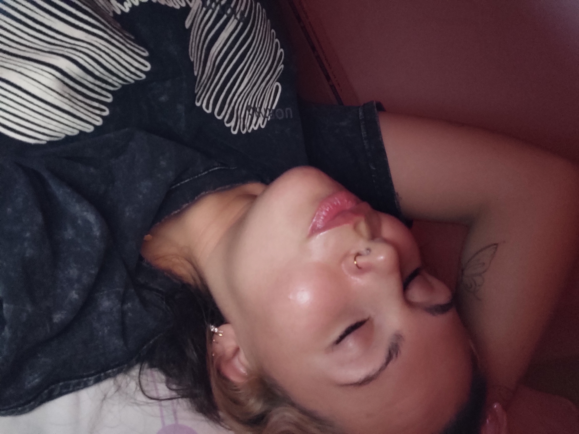 An intimate, close-up photograph captures a person, likely a young woman, in a state of rest or sleep, lying horizontally on a light-colored surface, possibly a bed, indoors in Ольховка, Russia. The warm-toned image shows her head turned slightly to the left, with her eyes closed in peaceful repose. Her fair skin appears smooth with a slight, natural sheen, highlighting her cheekbones, forehead, and nose. Her lips are slightly parted and coated with a glossy, pink balm or lipstick. A prominent gold septum piercing adorns her nose, and a small, delicate earring is visible on her left earlobe. Dark hair, possibly with lighter streaks or highlights, frames her face and spreads around her head. She is wearing a dark, possibly charcoal-colored, t-shirt featuring an abstract pattern of bold white wavy lines. A partial word "on" can be faintly seen printed on the shirt. Her right arm is bent with her hand behind her head, revealing a delicate, outlined butterfly tattoo on her upper arm. The background behind her is a soft, muted reddish-pink, suggesting a wall or soft ambient lighting that contributes to the image's overall warm atmosphere.