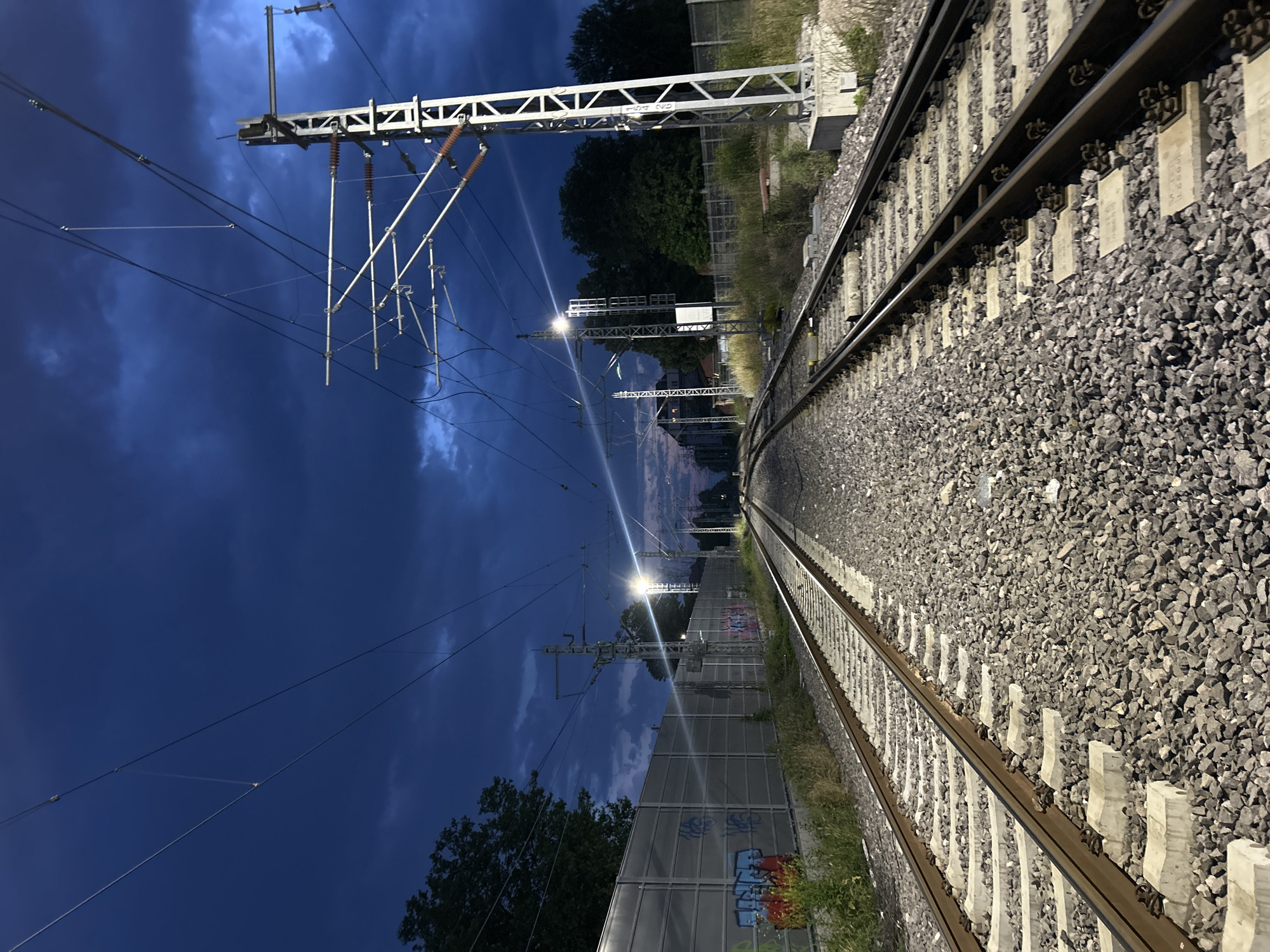 The vertically oriented image captures a night-time or late-dusk view of a dual-track electrified railway line. The tracks run diagonally from the bottom-right towards the middle-left, curving gently as they recede into the distance. They are composed of dark metal rails, supported by light grey concrete sleepers, all set within a bed of dark grey gravel ballast.
To the left of the tracks and occupying a significant portion of the upper frame is the intricate overhead catenary system. This includes large metal support gantries, individual masts, numerous taut wires, and various insulators. Several bright artificial lights are integrated into these structures, illuminating the overhead lines and casting light onto the tracks and surrounding area.
The sky, visible mostly in the upper-left and central parts of the image, is a deep blue, characteristic of night, and features scattered clouds. Along the lower-left edge of the tracks, a tall, partially translucent noise barrier or fence is present. This barrier is covered in colorful graffiti, displaying letters and shapes in red, blue, green, and black. Beyond the barrier and further down the line, dark trees and some distant, dimly lit buildings can be seen. On the right side of the tracks, sparse vegetation and trees line the embankment.
The scene is devoid of people or active trains, presenting a static view of railway infrastructure. Faint alphanumeric codes, such as "103 001", are visible on white plates attached to the overhead line support structures. Some concrete sleepers also bear indistinct text, likely identification marks. The image portrays a quiet, industrially lit atmosphere.