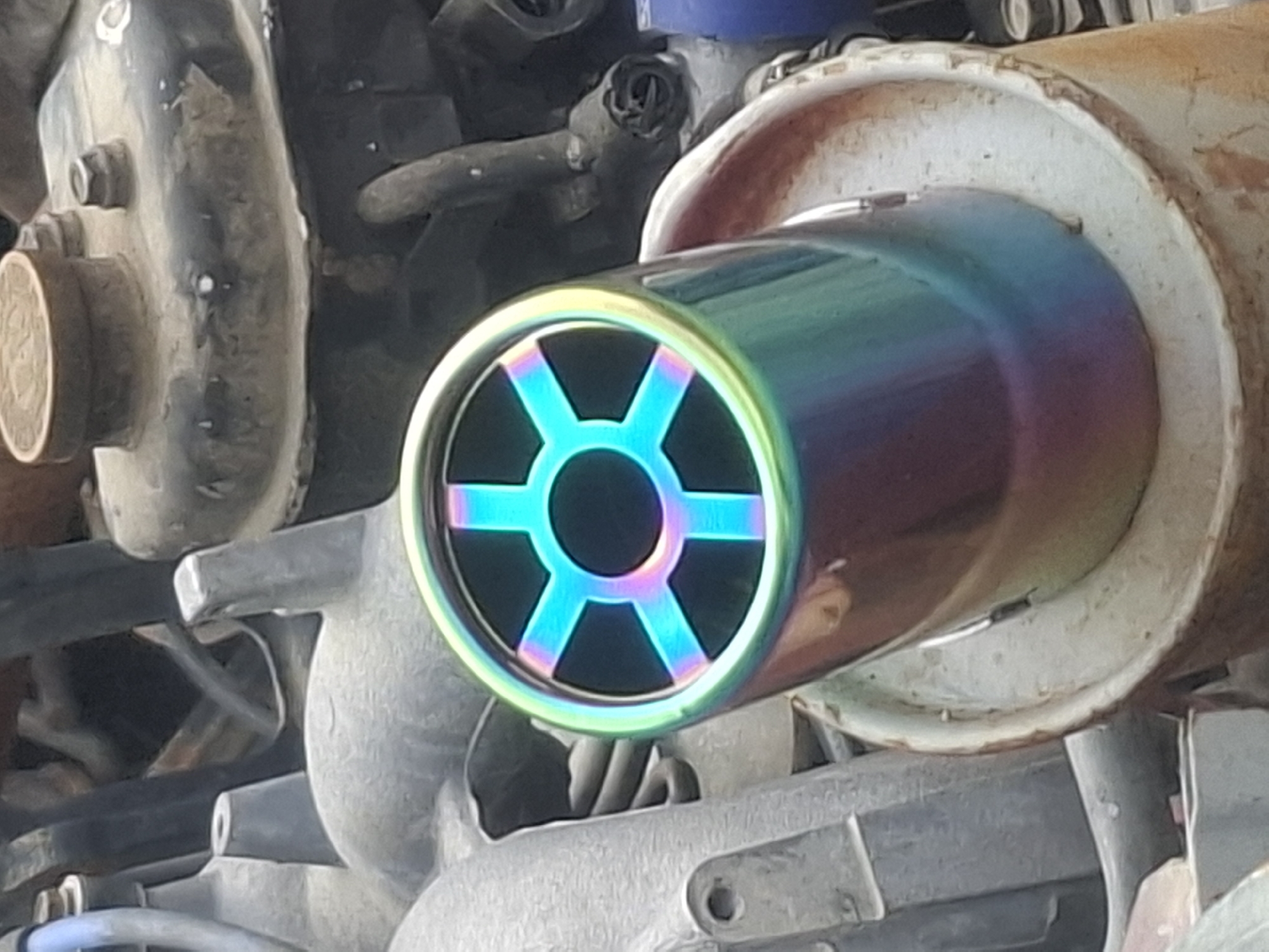 The image shows a close-up, low-angle view of a car's exhaust pipe with a rainbow-colored, perforated tip. The exhaust tip has a hexagonal pattern of spokes within a circular opening, illuminated by bright cyan and pink light. The exhaust pipe itself is cylindrical and exhibits a gradient of iridescent colors, transitioning from blue and green to purple and red. The exhaust pipe is inserted into a white, ceramic-like mounting. In the background, various grey, metallic engine components are visible, including hoses, pipes, and what appears to be part of the engine block. The overall environment suggests an automotive setting, likely a garage or a repair shop, or possibly a street view in Almaty, Kazakhstan, given the context provided. There are no people or discernible actions captured in this static image.