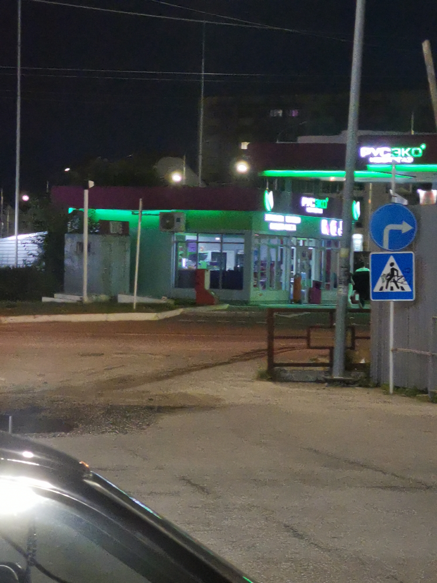 At night, a street scene unfolds in Cherkessk, Russia. A modern building with a purple facade and bright green neon lighting features a storefront with large windows. A sign with "РУСЭКО" in glowing white letters is visible on the building. Two road signs are posted: a blue circular sign with a white right-pointing arrow and a blue square sign with a white triangle containing a silhouette of a person walking across a road. A dark-colored car is partially visible in the foreground, its headlights reflecting on the wet asphalt. The environment is dimly lit, with the building's lights being the primary illumination.