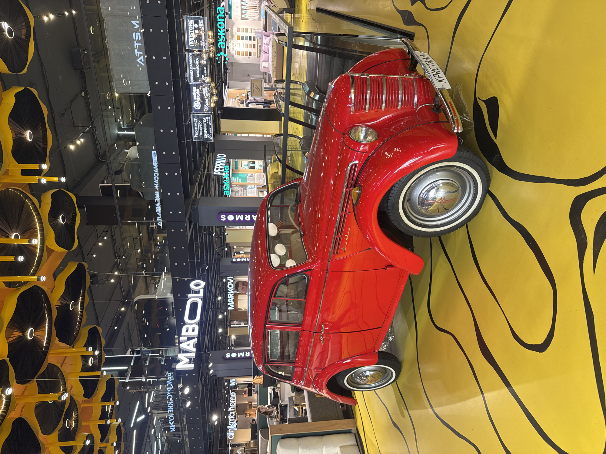 Captured indoors, the photograph features a classic red car mounted on a yellow floor with black, organic lines. The car is a focal point, its vintage design contrasting with the modern backdrop. Surrounding the car are various storefronts, marked with signs such as "MA'BOLO" and "MARKOVI," suggesting the car may be located within a shopping center. The lighting is bright, illuminating the scene and highlighting the colors. Yellow, wheel-shaped light fixtures hang from the ceiling on the left. The image appears to be taken in a public space, likely within a retail or commercial setting. The license plate is "КРИСТАЛЛ".