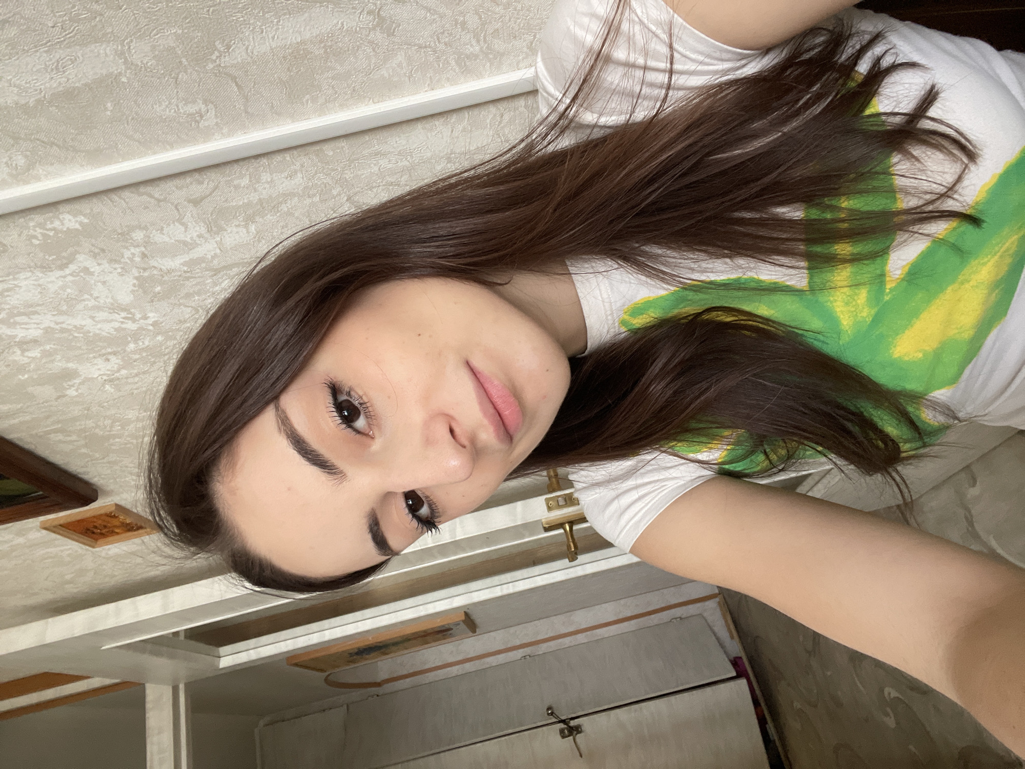 A selfie of a woman with long brown hair wearing a white t-shirt with a green and yellow design. The background includes a white ceiling with textured wallpaper, interior architectural details, and a portion of a light-colored door or cabinet with brass hardware. There is no evidence indicating the location is Moscow, Russia.